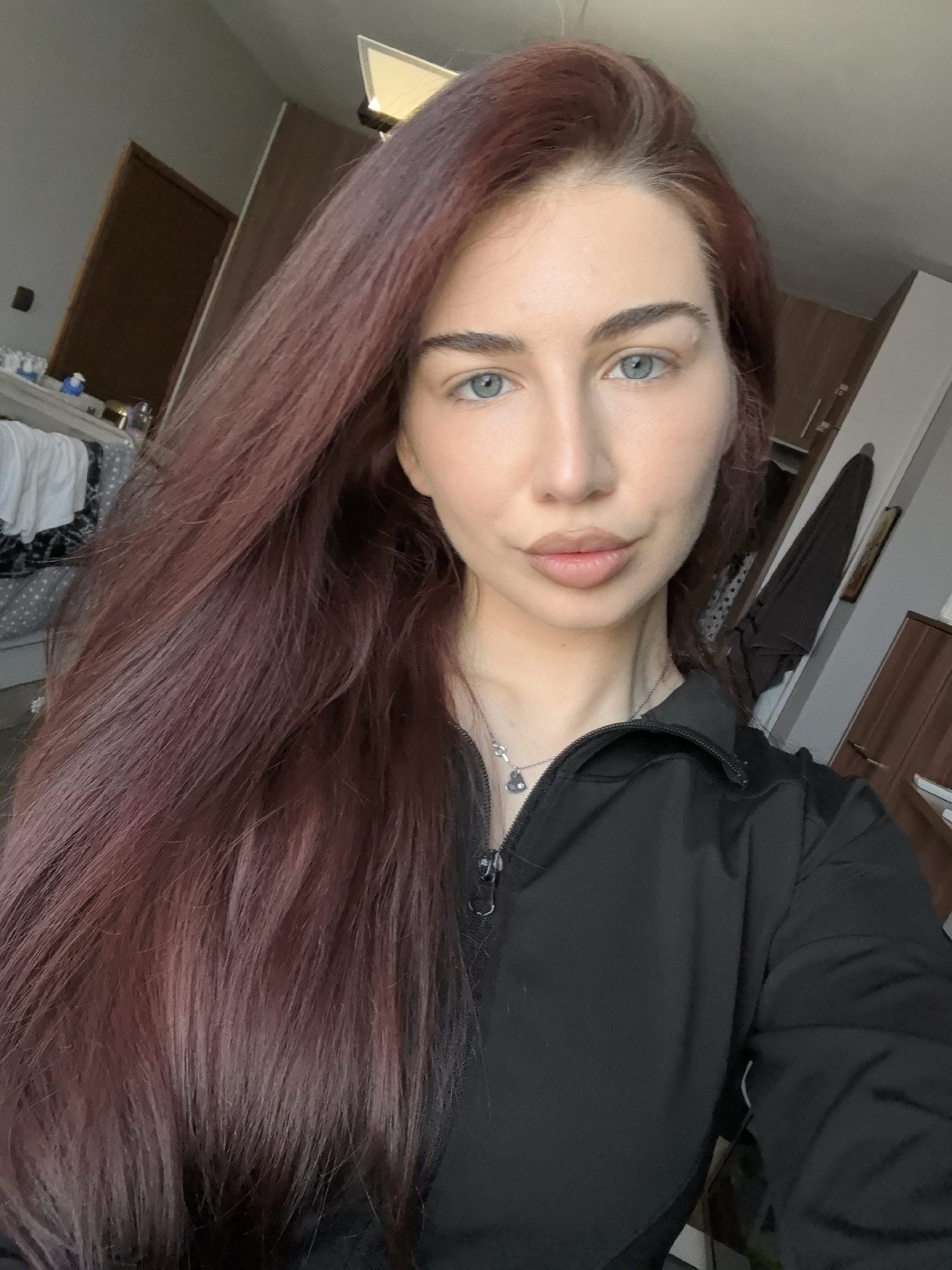 A woman with long, auburn-red hair poses indoors. Her eyes are light blue. She wears a black zippered jacket and a silver necklace. The background shows a bedroom with a partially visible bed, white linens, and a dark wooden dresser. There's a door and a hanging coat visible to the right. There are no specific indicators confirming the location is Stara Zagora, Bulgaria.