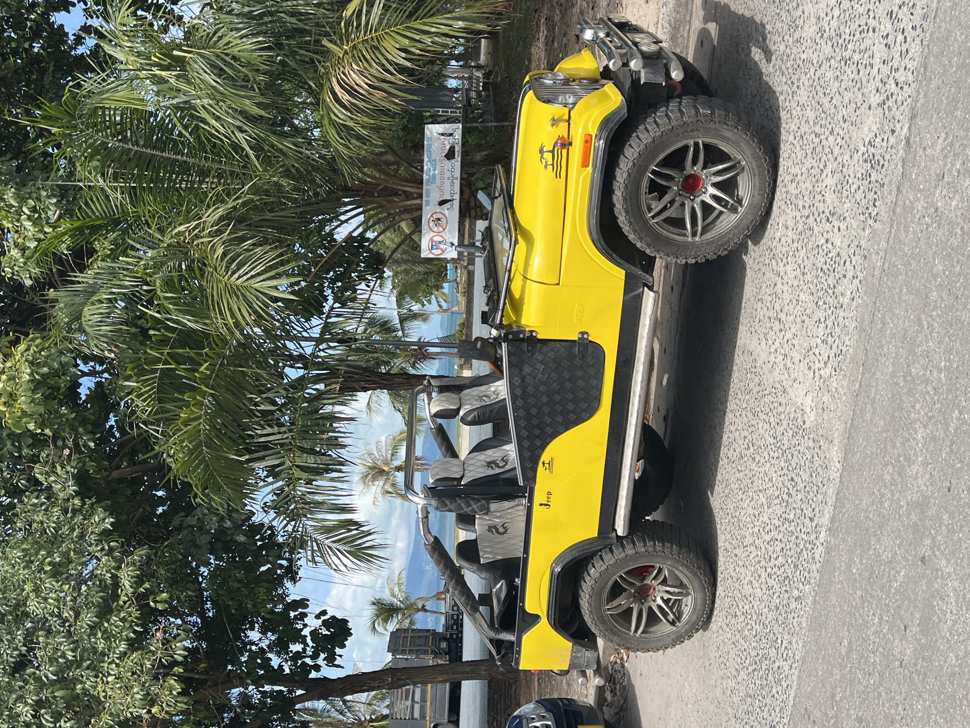 A bright yellow, open-top Jeep is parked on a paved road. The Jeep has custom silver wheels with red accents and black accents on the sides. The interior features black seats with silver diamond-patterned upholstery and dragon graphics. A sign with international symbols and text in Thai and English is visible in the background. Behind the Jeep, palm trees and other foliage are abundant, with a hint of the ocean and mountains in the distance. The sky is blue with scattered clouds. The scene appears to be in a tropical coastal area.