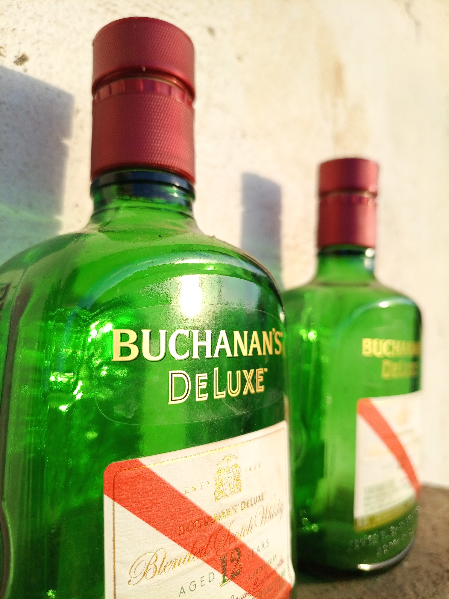 The image shows two green glass bottles of Buchanan's Deluxe Scotch Whisky, with red caps. The label on the bottle in the foreground features a diagonal red stripe across a white background, and the words "Buchanan's Deluxe" and "Blended Scotch Whisky". The bottle in the background is slightly out of focus, but shows a similar label. The location is Unknown, Unknown.