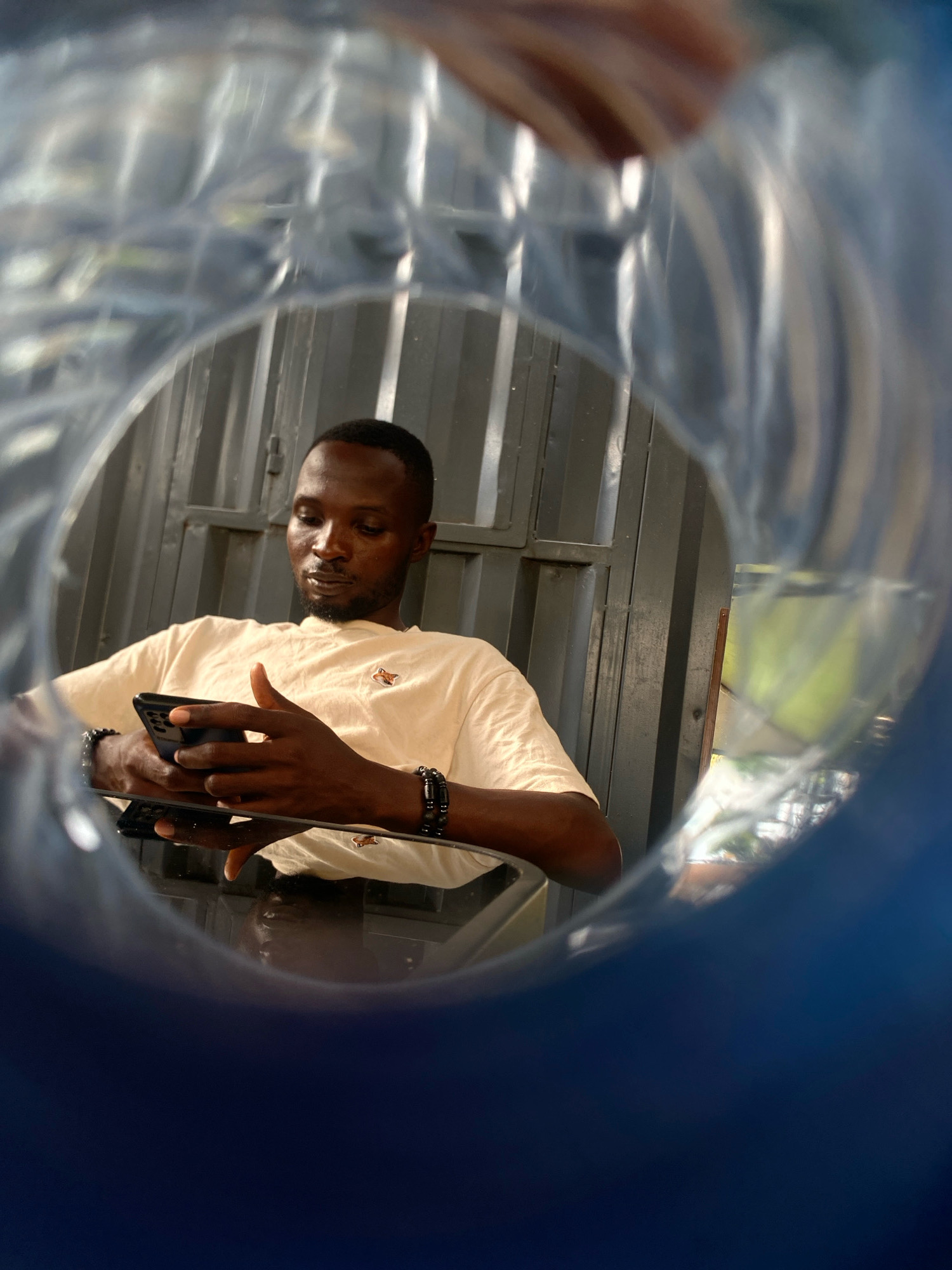 A man is seen from inside a blue plastic bottle, looking down at his phone. He is wearing a cream-colored t-shirt with a small fox logo and two black beaded bracelets. The background is a corrugated metal wall. The lighting suggests it is daytime. The image was taken in Barden Yana, Nigeria.