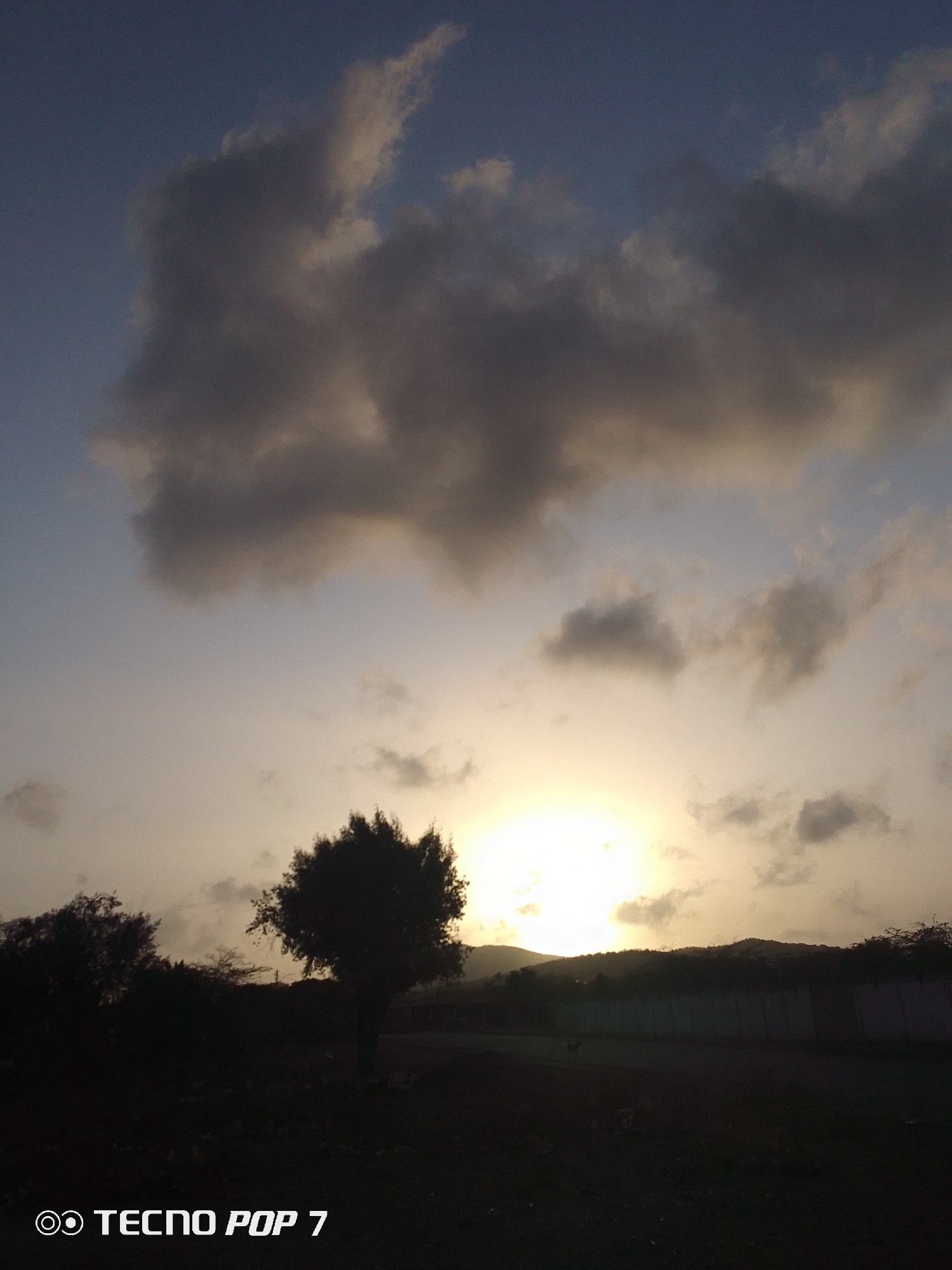 The image captures a sunset scene in Altagracia, Venezuela. Dominating the upper portion are large, dark clouds against a sky gradient of blue and yellow, indicating the sun's position. The sun is obscured by the horizon, creating a bright glow. In the foreground, silhouetted trees stand against the horizon. A long, low wall runs horizontally across the middle ground. The bottom portion is mostly dark, obscuring details. The text "TECNO POP 7" is visible in the lower left corner, suggesting the camera used to take the picture.