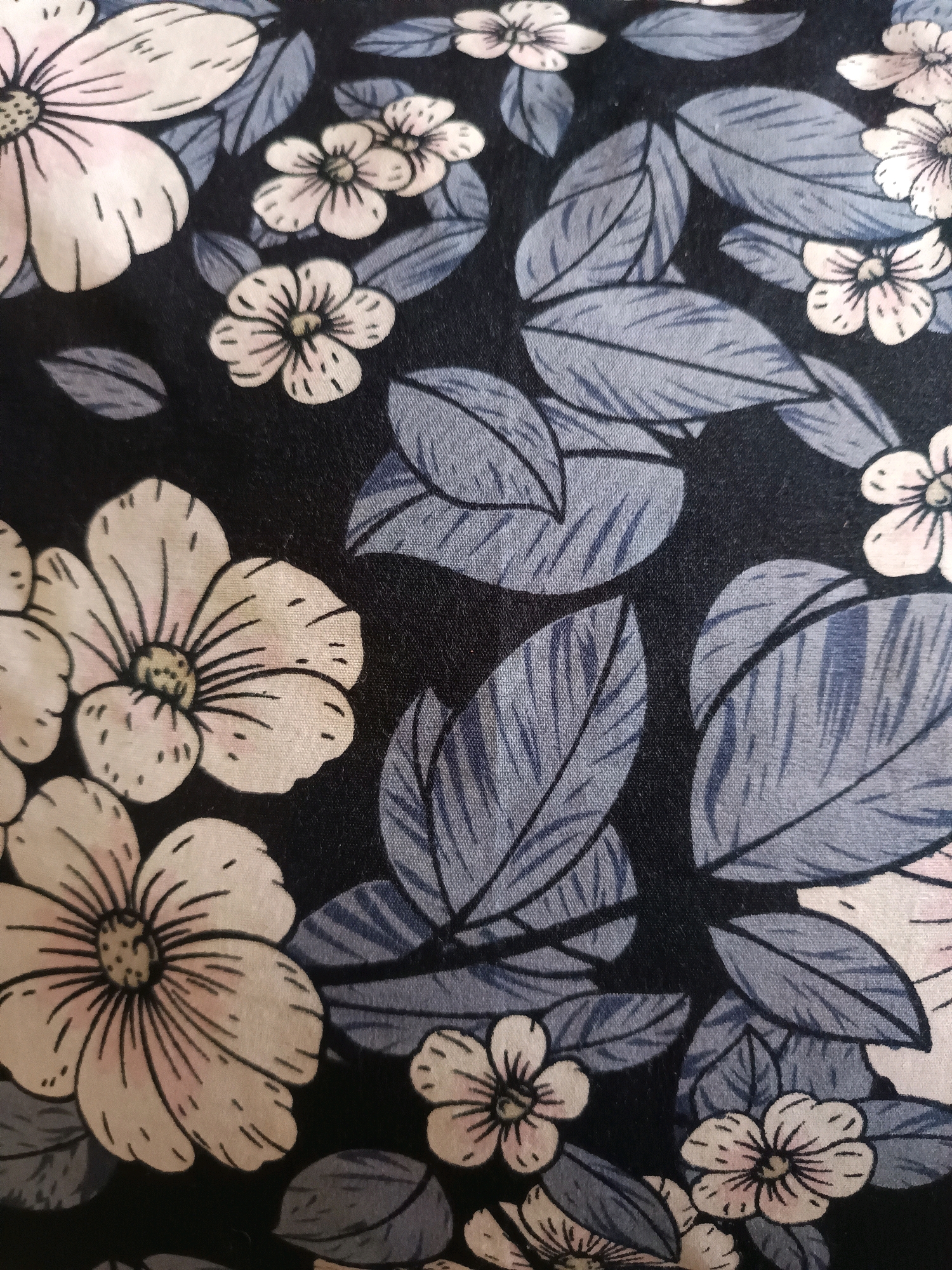 The image shows a close-up of a dark fabric with a floral pattern. The pattern features light pink and white flowers with black outlines, interspersed with blue-grey leaves. The flowers have multiple petals and visible stamens in the center, with some petals showing delicate pink shading. The leaves are elongated and veined. The background is a solid dark color, likely black, which makes the floral and leaf elements stand out. The texture of the fabric is visible, suggesting it might be a woven material. There is no discernible urban environment or specific location mentioned within the image itself, therefore it cannot be placed in Gunung Sekar, Indonesia.
