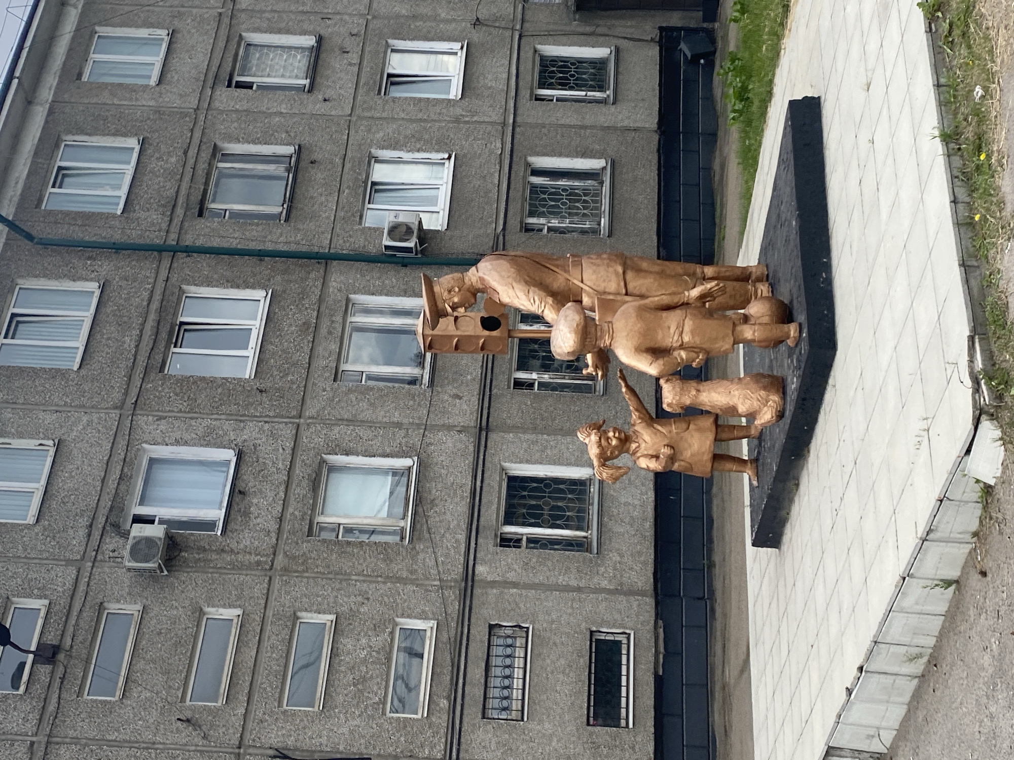 A golden bronze sculpture depicts a man in a postal uniform, possibly a postman, standing next to a traffic light signal that appears to be a part of the sculpture. He is interacting with two children and a dog, all also rendered in golden bronze. The scene is set against a backdrop of a gray, multi-story apartment building with numerous windows and air conditioning units. The building's facade is textured and appears to be made of concrete panels. The sculpture is situated on a raised concrete platform paved with square tiles, surrounded by a low, damaged concrete curb and overgrown grass. The overall setting suggests an urban environment in Usolye-Sibirskoye, Russia. The children are positioned at the man's feet, one reaching out towards him, while the dog sits beside them. The man appears to be addressing the children, and the traffic light is positioned behind him. The image is taken from a low angle, emphasizing the sculpture and the building.