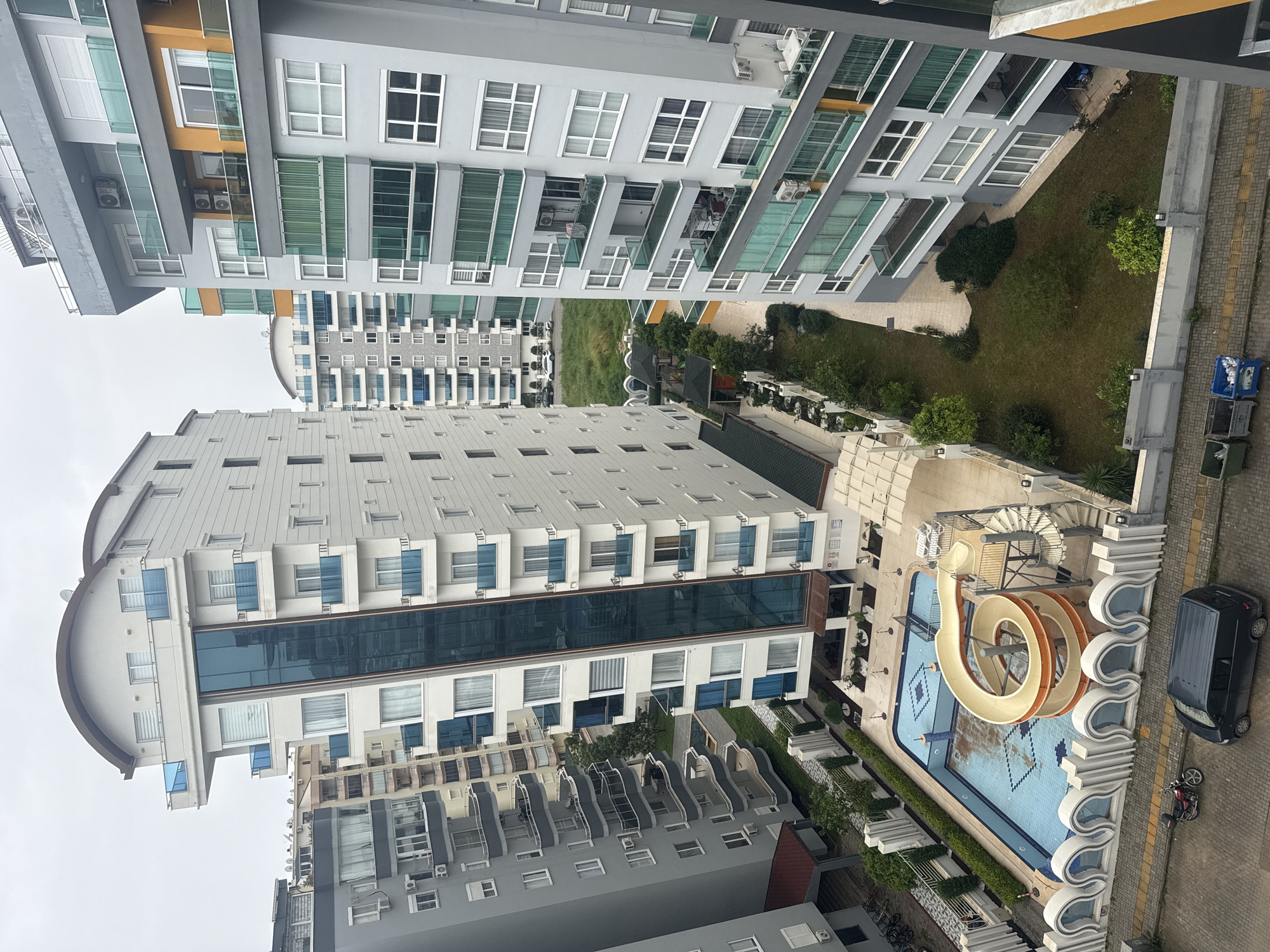 The image depicts a high-angle view of an urban landscape in an unknown city. The scene is dominated by several multi-story apartment buildings with modern architectural designs. A large, curved swimming pool with a water slide is visible near the bottom of the image, adjacent to a building. A black van and a parked motorcycle are near the pool. Between the buildings are patches of greenery, including grass and trees. The overall atmosphere is residential and suggests a densely populated area. The weather appears overcast.