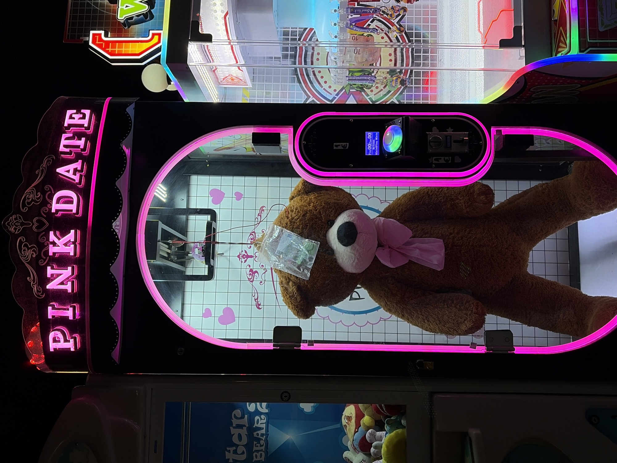 A large, brown, plush teddy bear with a pink bow is displayed prominently inside a brightly lit arcade machine. The machine features a pink neon outline and signage that reads "PINK DATE" in large, illuminated letters at the top. The bear has a plastic bag taped to its forehead, containing small items. The background of the machine is a tiled wall with pink heart decals. To the left of the "PINK DATE" machine is another arcade machine with blue and white graphics. To the right, other arcade machines are visible, some with colorful displays and prizes. The scene is illuminated by the neon lights of the machines, suggesting it is nighttime or an indoor, dimly lit environment. The location is identified as Boreti, Montenegro.