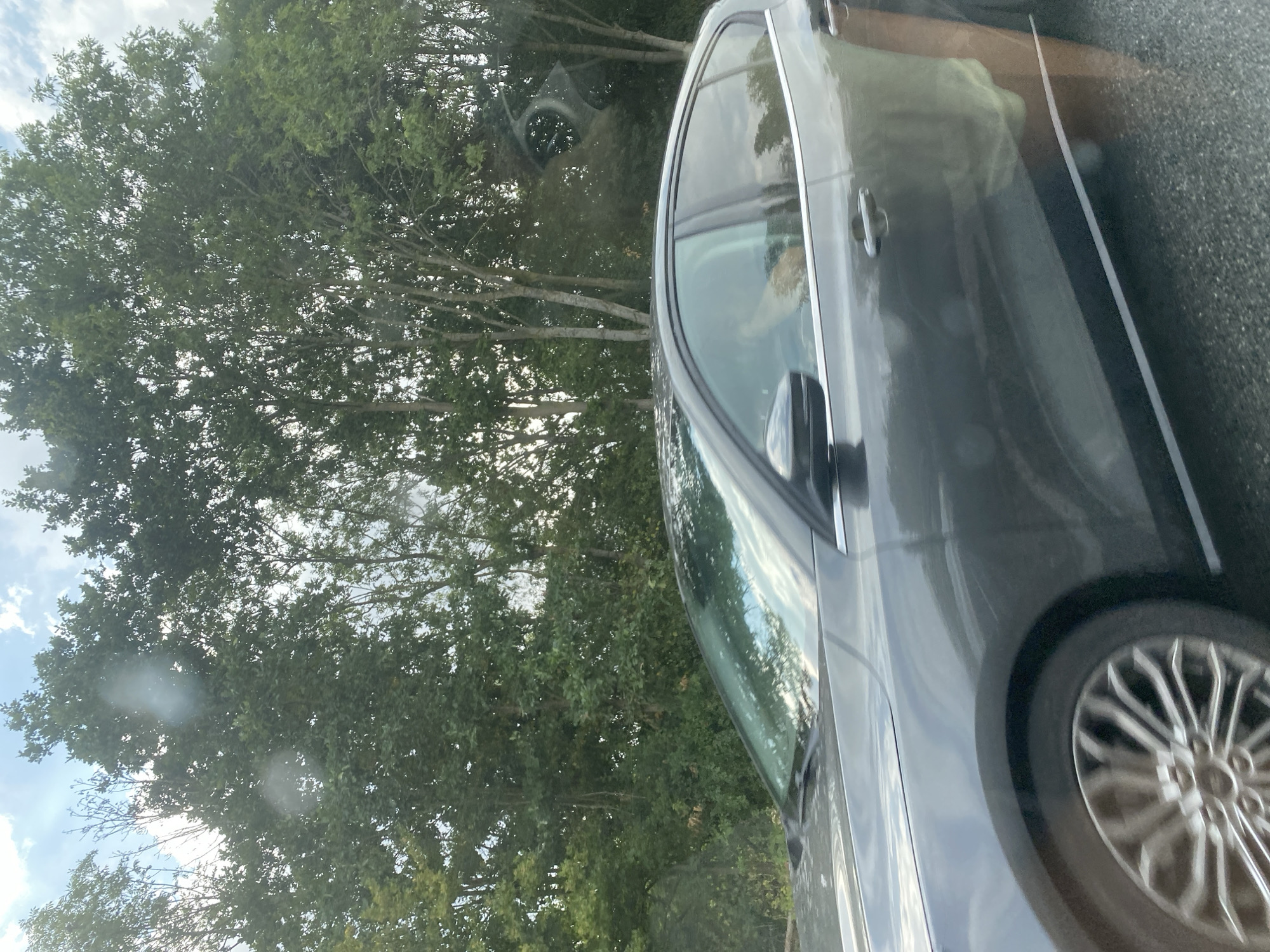The image shows a dark gray sedan driving on a road with trees lining the background. The sky is partly cloudy. Inside the car, a person's hand can be seen resting on the center console, suggesting they are the driver or a passenger. The car appears to be in motion, as indicated by the blur on the wheel and the overall impression of movement. The scene is likely captured from another moving vehicle. The location is Weyhe, Germany, a municipality in the district of Diepholz, Lower Saxony.