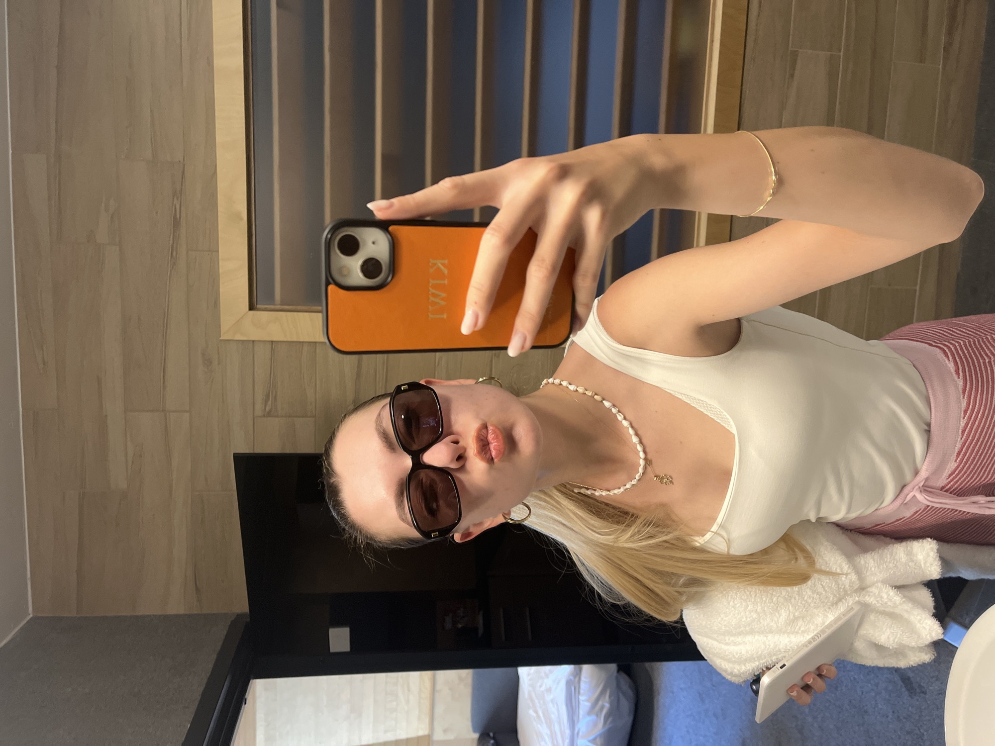 A woman takes a selfie in a hotel bathroom. She is wearing sunglasses, a white tank top, a pearl necklace, and a bracelet. She is holding an orange phone case with her name on it. The bathroom has a tiled wall, a TV, a bed with white sheets, and a white towel.