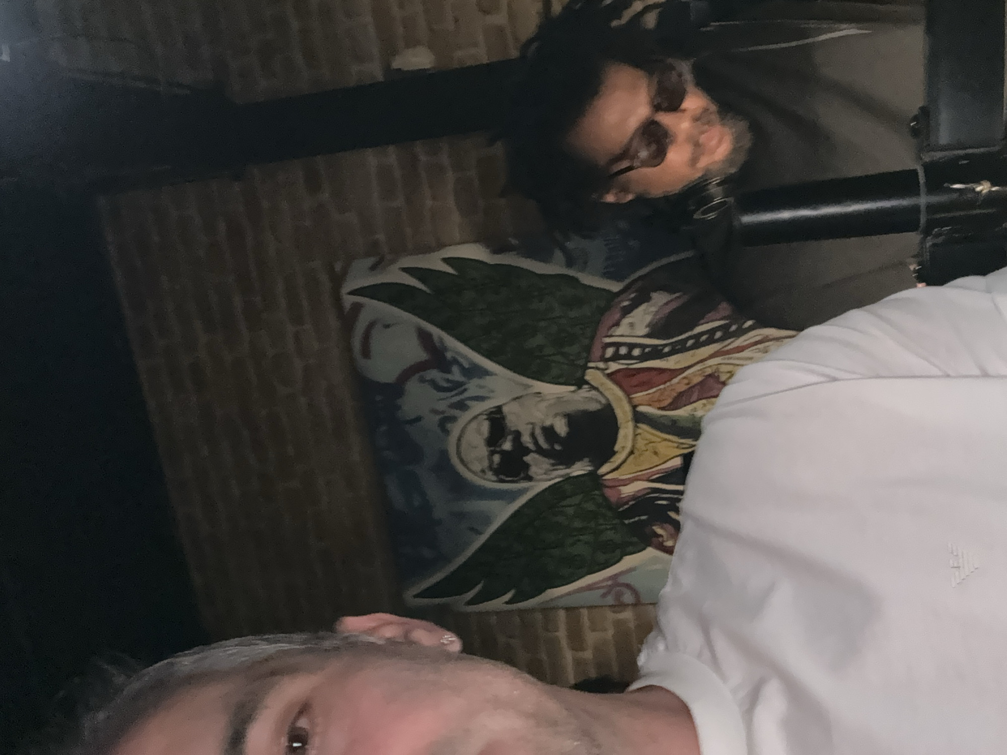 A selfie taken from a low angle shows two individuals in an indoor setting with a brick wall and a painting. In the foreground, the left side of a person's face and upper body are visible, wearing a white t-shirt with a small logo. The person's skin is light, with short grey hair and an earring. To the right and in the background, a second person is visible, appearing to be wearing sunglasses and a dark-colored top. This individual has dark, curly hair and is leaning forward. Behind them, a painting on the wall depicts a person with wings, possibly a stylized portrait. The lighting is dim, suggesting an evening or indoor event. The overall impression is a casual, social gathering.