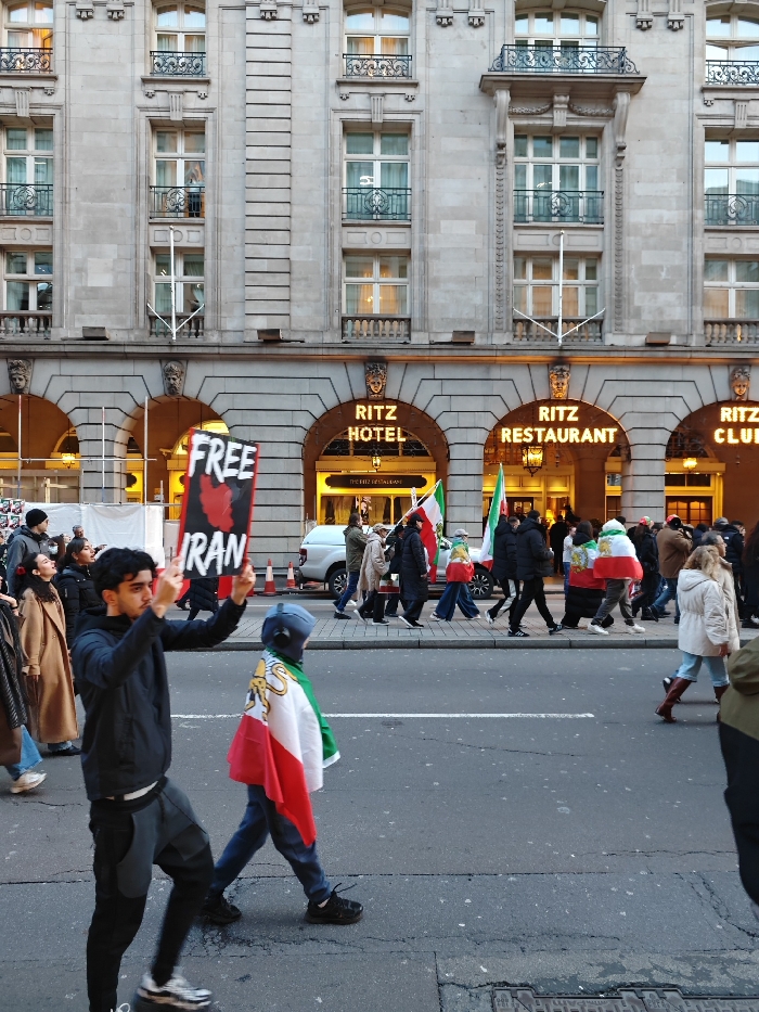 The image depicts an outdoor street scene in what appears to be late afternoon or early evening, characterized by both natural light and activated artificial lighting from buildings. The focal point is a public demonstration or protest taking place in front of a grand, ornate building identified as The Ritz London.
In the background, the multi-story stone facade of The Ritz Hotel stands prominently. Its classical architecture is evident with numerous windows, some featuring decorative balconies and balustrades. Above the ornate arched entrances, illuminated signs clearly read "RITZ HOTEL," "RITZ RESTAURANT," and "RITZ CLUB" in golden lettering, indicating different sections of the establishment. Lights are visible inside the hotel and restaurant, illuminating the entrances.
In the foreground and midground, a diverse group of people is moving along the street and sidewalk, seemingly in a procession. Many participants are carrying or draped in flags that resemble the historical flag of Iran, typically featuring green, white, and red horizontal stripes with a golden lion and sun (Shir-o-Khorshid) emblem in the center.
A young man in the immediate foreground, wearing a dark jacket and dark pants, is holding a black sign high above his head with both hands. The sign bears a clear message in white and red text: "FREE IRAN." The word "FREE" is in white, and below it, the word "IRAN" is also in white, set against a red background that seems to include a stylized symbol, possibly a fist or another protest graphic. He is looking towards the left side of the frame.
To his right and slightly behind him, another individual, possibly a child or young person, is walking with an Iranian flag draped over their shoulders, its colors and the lion and sun emblem visible. This person is wearing a blue or grey hooded garment.
Further down the street, a larger group of demonstrators continues to walk, with more Iranian flags visible among them. A white van is parked on the street near the Ritz entrance. The ground is a paved road with visible road markings and a concrete sidewalk. The scene suggests a public gathering expressing solidarity or protest concerning Iran.