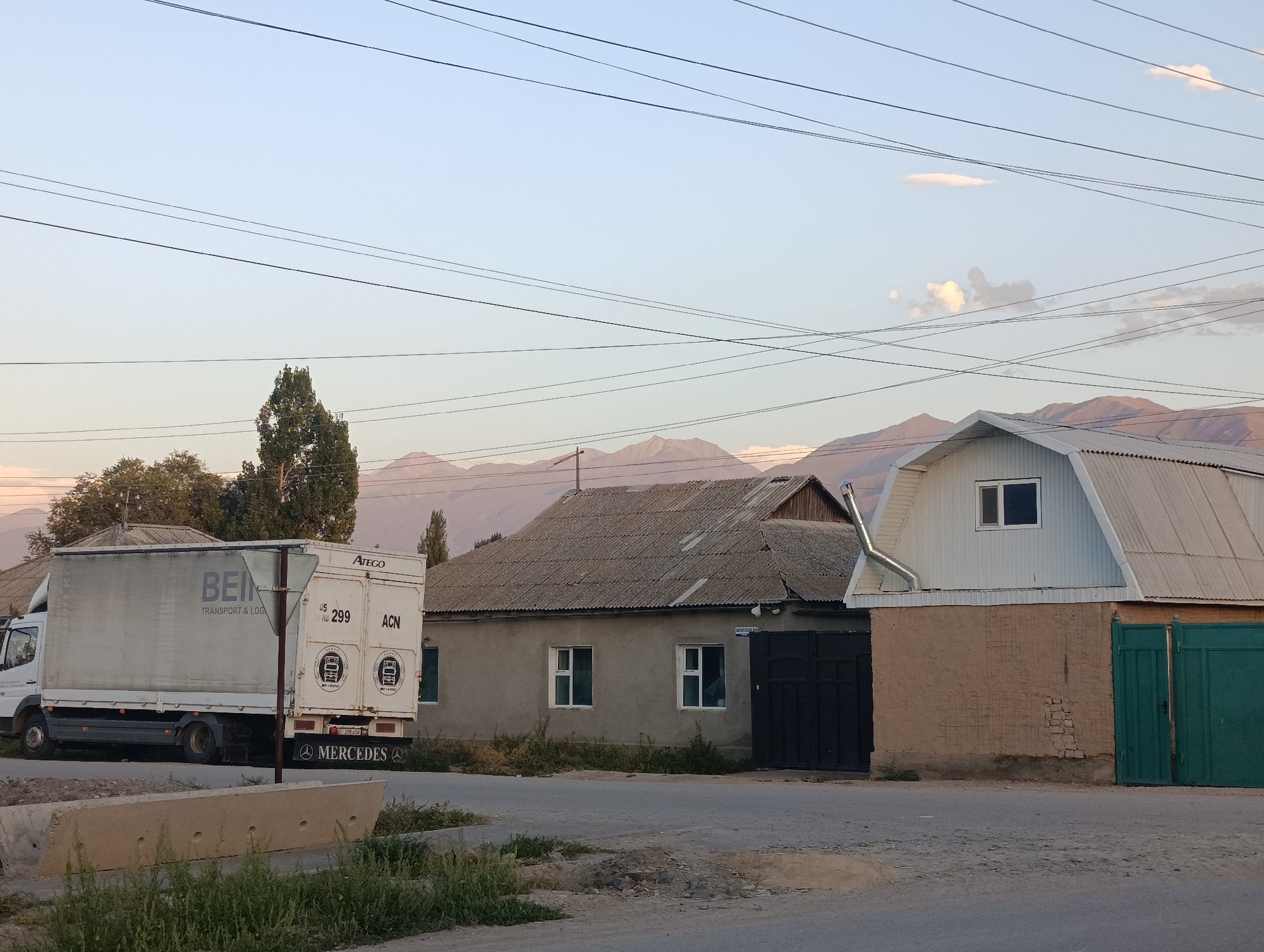 In Kochkor, Kyrgyzstan, a street scene unfolds under a pale blue sky with scattered clouds and distant mountains. A white Mercedes-Benz Atego truck, a commercial vehicle with a large cargo box, is parked on the left side of the image. The cargo box is mostly obscured by the truck's cab, but a portion of the side is visible with branding that includes "BEIN TRANSPORT & LOGISTICS."
To the right of the truck, a single-story, weathered building with a corrugated roof is visible. It features two windows with white frames and a dark, large double gate on the right side. Next to this building is another structure with a light-colored, gabled roof, also with a corrugated metal finish. This building has a small window on its upper level and a section of exposed brickwork. A vibrant green metal gate is situated to the far right.
The foreground shows a paved road with a concrete barrier and patches of dry grass and weeds. The overall impression is one of a rural or semi-urban environment in Kyrgyzstan, likely captured during the late afternoon or early evening given the lighting. The presence of overhead power lines crisscrossing the sky adds to the utilitarian feel of the scene.