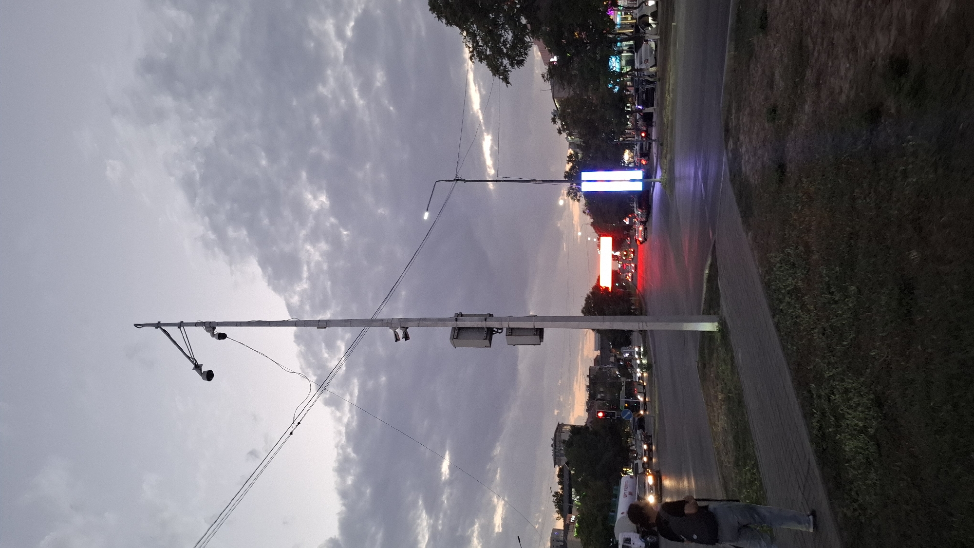 The image depicts a street scene in Shymkent, Kazakhstan, during twilight. A tall pole with multiple surveillance cameras and traffic signal equipment stands prominently in the foreground, positioned near a sidewalk and grassy area. The street is active with vehicular traffic, illuminated by headlights and traffic signals, including red and blue lights from digital signs. In the background, buildings and trees are visible, silhouetted against a cloudy sky with hints of sunset colors. A person with a backpack is walking on the sidewalk, facing away from the camera. The overall atmosphere suggests a typical urban environment at dusk.