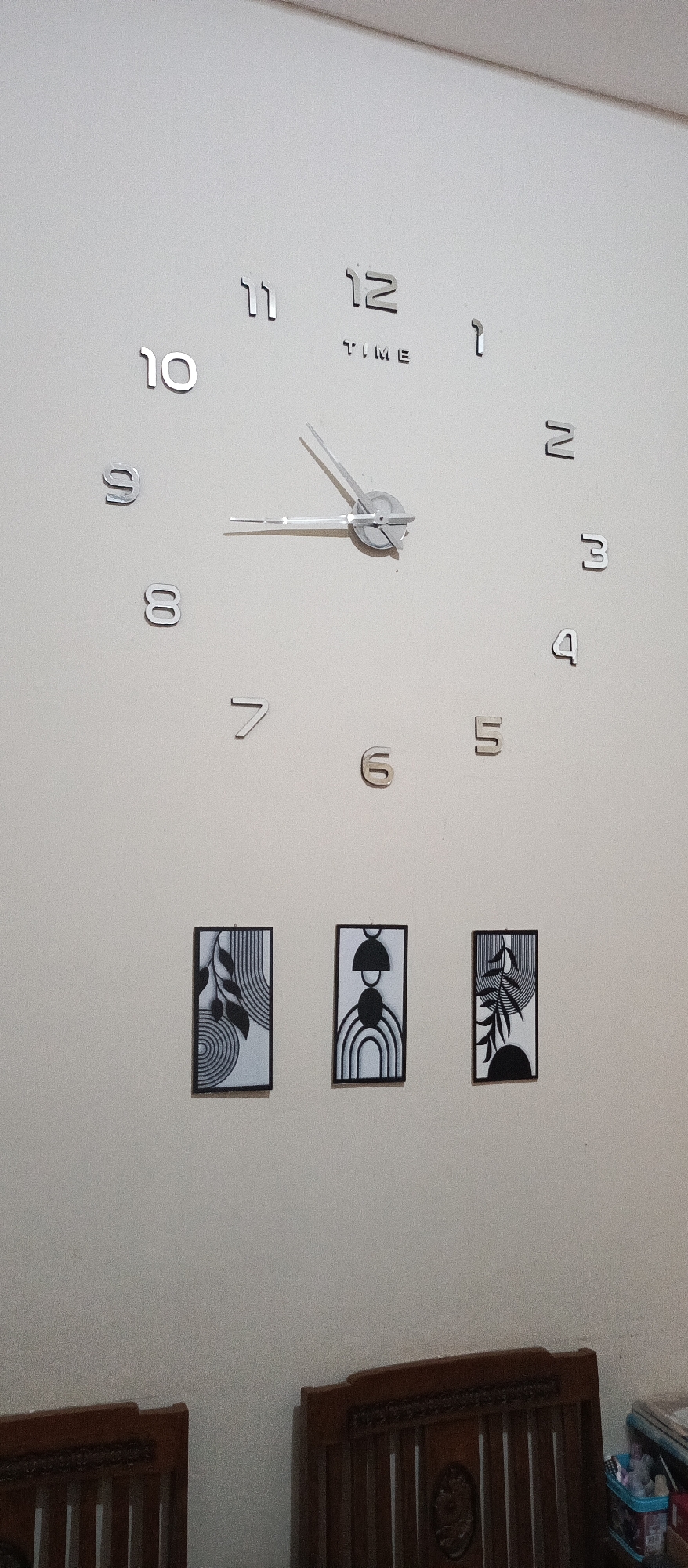 The image shows a large, modern wall clock with silver numbers and metallic hands mounted on a plain light-colored wall. The clock hands indicate the time is approximately 10:10. Below the clock, three framed abstract art pieces with black and white designs are displayed. The art pieces are arranged horizontally. In the foreground, the tops of two wooden chairs or benches are visible, suggesting an indoor domestic setting. The overall lighting is soft and diffused. The location is the living room of a house in Arjasa, Indonesia.