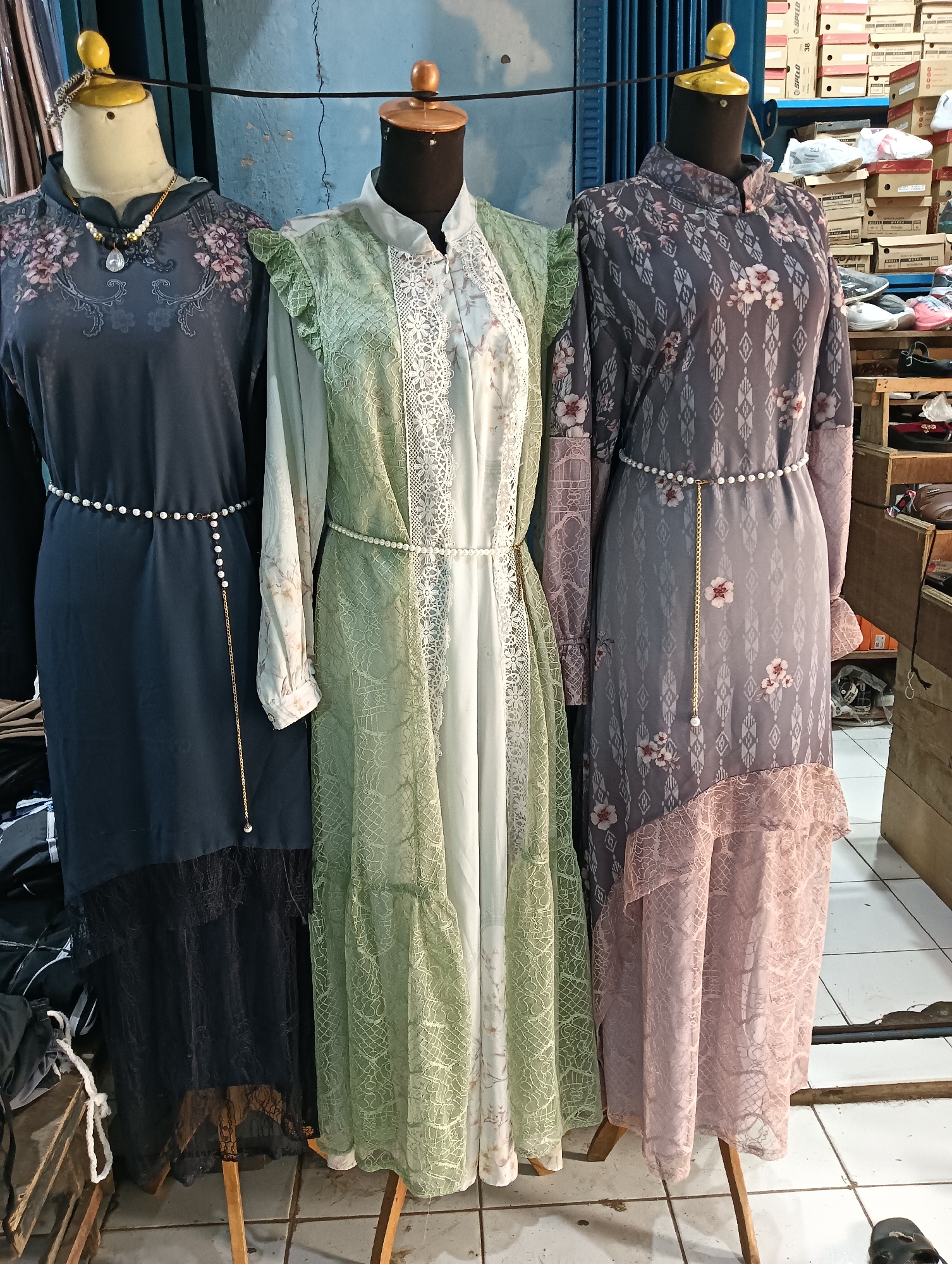 Three mannequins are displayed in what appears to be a shop in Cluwak, Indonesia, with clothing racks and shelves visible in the background. The mannequins are dressed in long, elegant dresses. The mannequin on the left wears a dark grey dress with floral embroidery on the shoulders and a black lace trim at the hem. It is accessorized with a pearl necklace. The middle mannequin is adorned in a light green dress with ruffles on the sleeves and a lace trim down the front. The mannequin on the right is dressed in a grey patterned dress with pink floral accents and a pink lace hem. All three dresses are cinched at the waist with pearl belts. The background shows stacks of shoeboxes and various items on shelves, suggesting a retail environment. The floor is tiled.