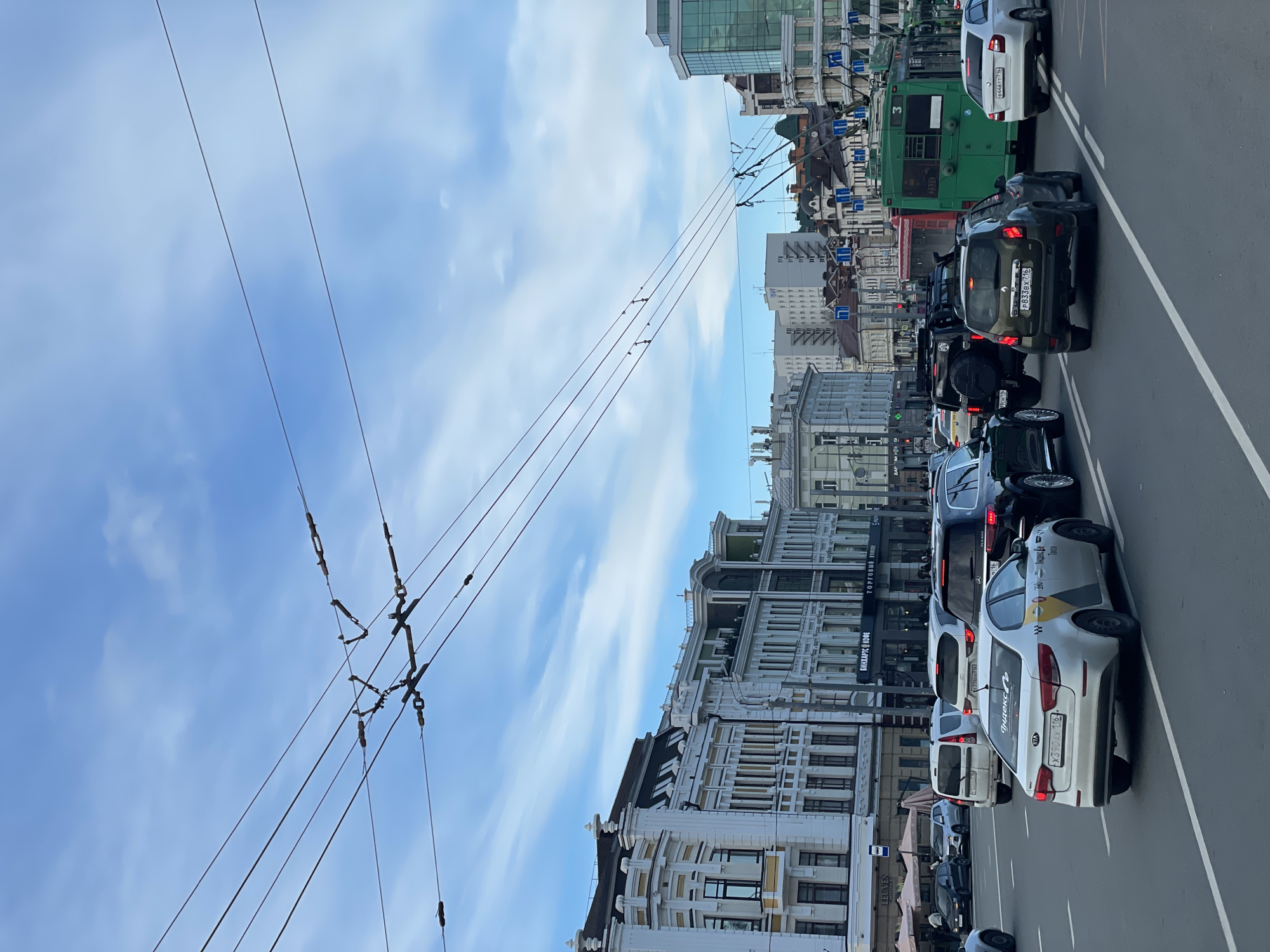 This image captures a busy city street in Kazan, Russia, seen during the daytime under a partly cloudy sky. The perspective is from a low angle, looking across a multi-lane road towards a row of diverse buildings.
A line of vehicles is queuing on the street, indicating typical urban traffic. These include a white Kia Rio taxi with "Яндекс.Такси" branding and the Russian license plate "X390AX 116", a dark SUV bearing the license plate "P833BX 716", a black Jeep-style vehicle, and a white sedan with the license plate "B446TT 716". A large green trolleybus, marked with route number "3" and vehicle number "2370", is also visible further down the road.
The buildings lining the street showcase a mix of architectural styles, from ornate, classical-looking structures with intricate details to more modern buildings featuring large glass panels. Overhead, a complex network of electrical wires for trolleybuses spans the sky, a common feature in many Russian cities.
Notable details include the bright, fair weather with a blue sky dotted with scattered clouds. Visible text on building signs includes "БИНХАРТС КОФЕ" (Binharts Coffee) and "ТОРГОВЫЙ" (Trade/Shopping), suggesting commercial establishments. The Russian language on the signs and the specific regional codes (116 and 716) on the license plates confirm the location as Kazan, Russia. The scene conveys an everyday urban atmosphere.