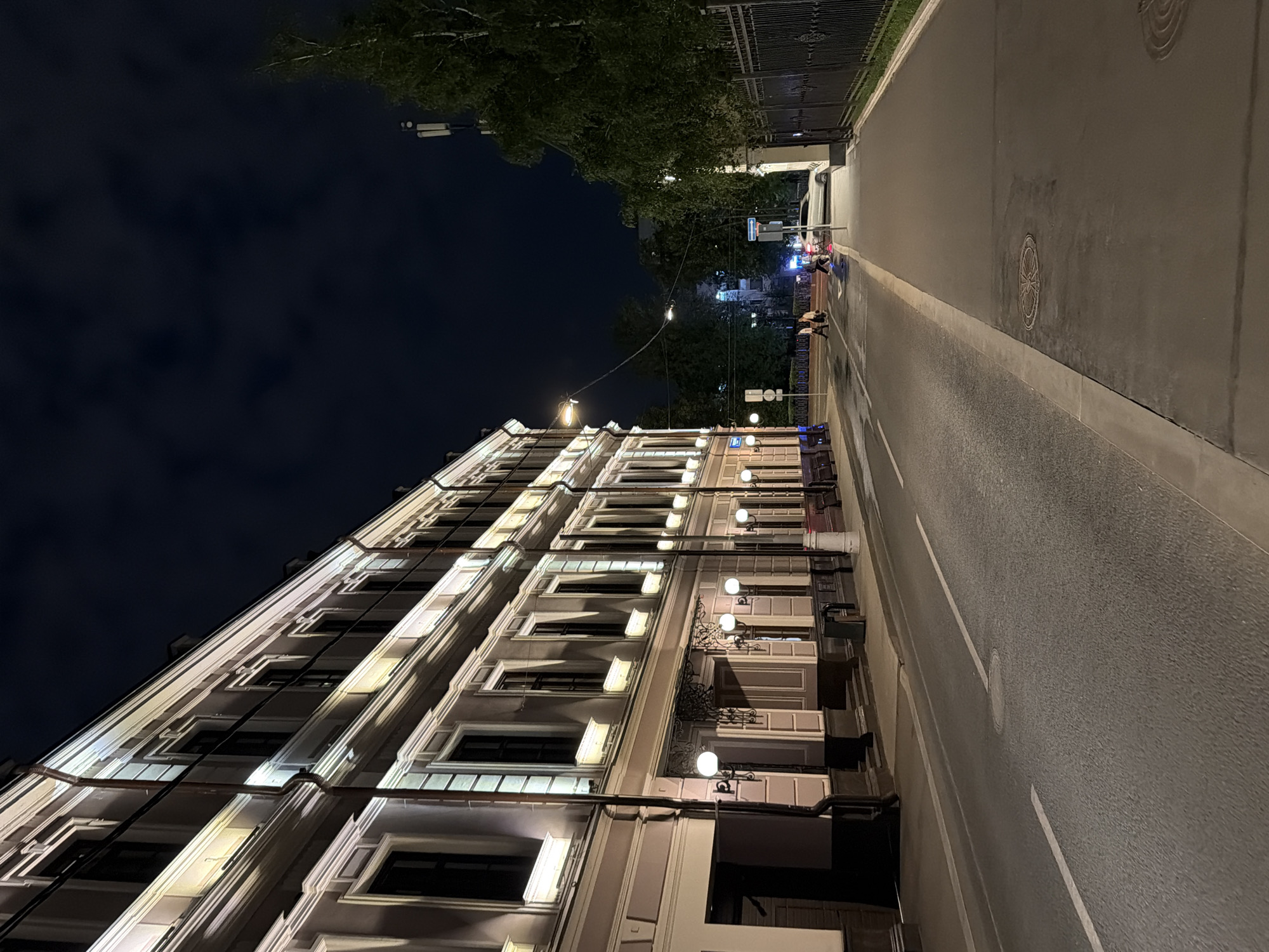 The image shows a street scene at night in Moscow, Russia. A long, multi-story building with illuminated windows and decorative facade lighting runs along the left side of the street. The building appears to be a historical structure with classical architectural elements. Across the street, there is a sidewalk with a black metal fence and lush green trees behind it. The road is asphalt, with white lane markings and a pedestrian crossing visible further down. A white car is driving on the right side of the road, and several people are walking on the sidewalk and crossing the street. The sky is dark with some clouds, and streetlights are visible, illuminating the scene. The overall impression is a quiet, urban night environment.