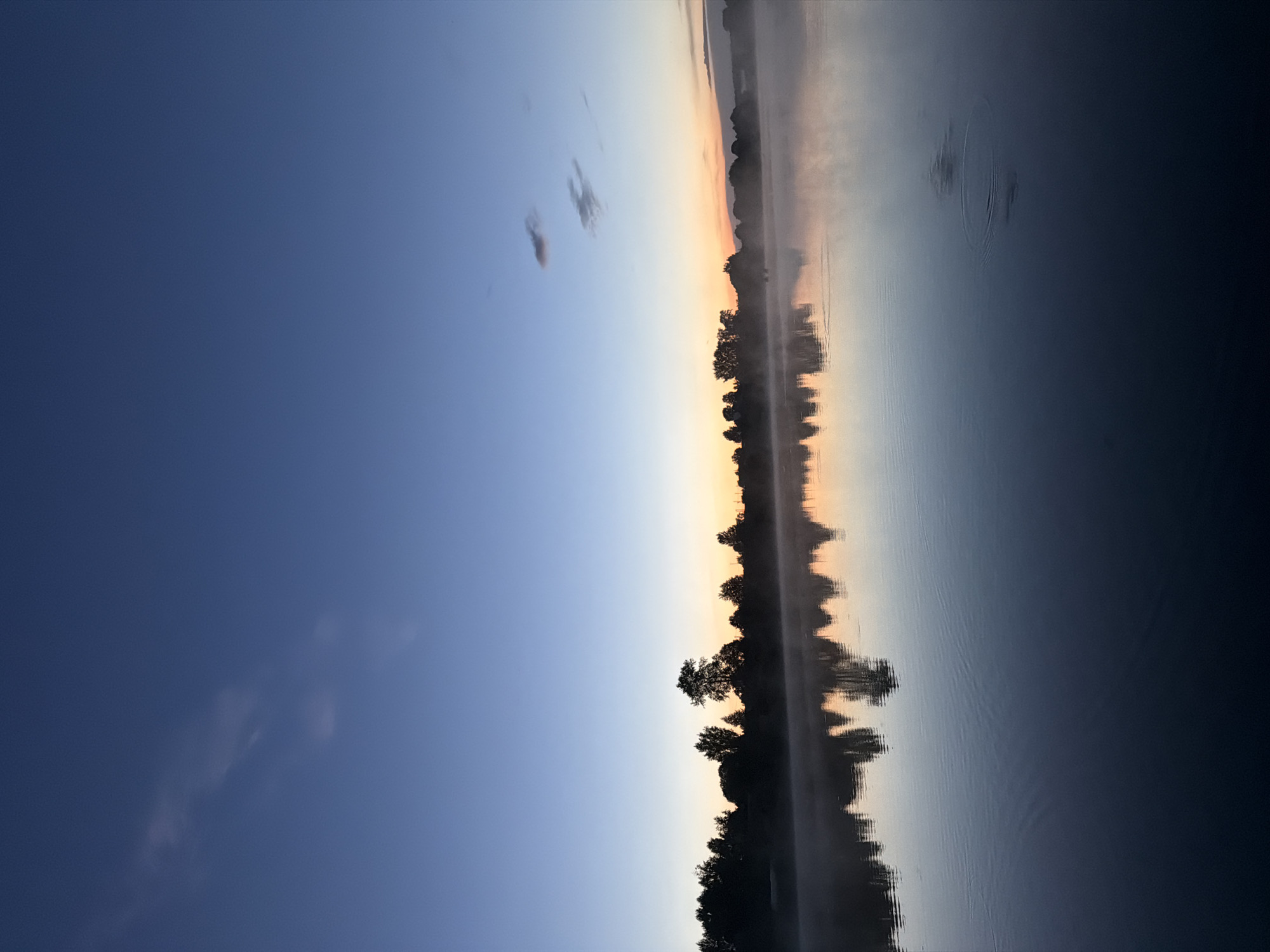 The image depicts a serene natural scene at dawn or dusk, likely a lake or wide river in Amaneevo, Russia. The sky transitions from a deep blue at the top to lighter shades of blue and orange near the horizon, indicating either sunrise or sunset. A thin line of trees silhouetted against the horizon forms the backdrop. A gentle mist hovers over the water, particularly along the shoreline, adding a soft, atmospheric quality. The water's surface is mostly calm, with subtle ripples and reflections of the trees and sky. There are no visible people or man-made structures, suggesting a secluded and tranquil natural environment.