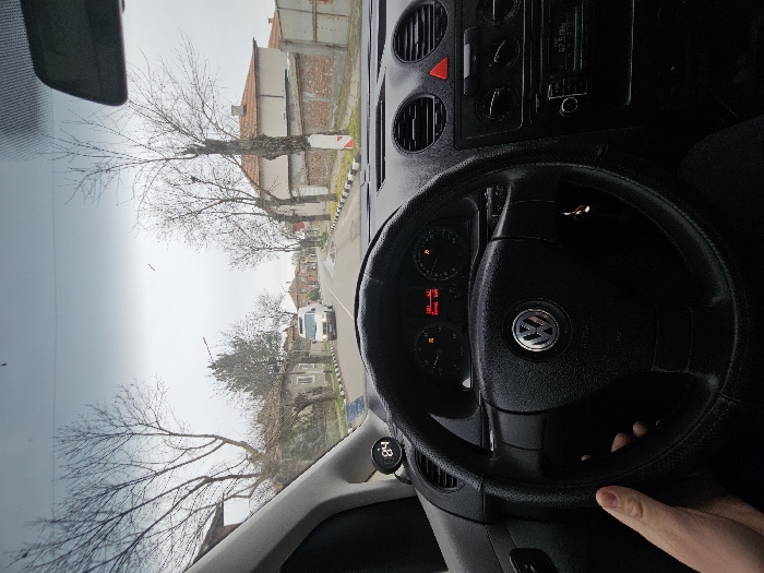 The image captures a first-person view from the driver's seat of a black Volkswagen vehicle on a street in Uzundjovo, Bulgaria, under an overcast sky. The driver's left hand is visible gripping the left side of the black, aftermarket-covered steering wheel, which prominently displays the Volkswagen logo. The dashboard's instrument cluster shows a digital time of "20:04" and a distance of "200 km", along with several illuminated warning lights. An auxiliary digital gauge displaying "84" is mounted on the far left of the dashboard. The center console features air vents, a red triangular hazard light button, climate controls, and a radio displaying "87.5 MHz".
Through the windshield, a paved road stretches into the distance, flanked by bare deciduous trees indicating a cold season. Residential buildings with tiled roofs and some brick fences line both sides of the street. A white van with its headlights on is visible approaching from the opposite direction in the middle distance. Black and white striped curb markings are present along the edge of the road.