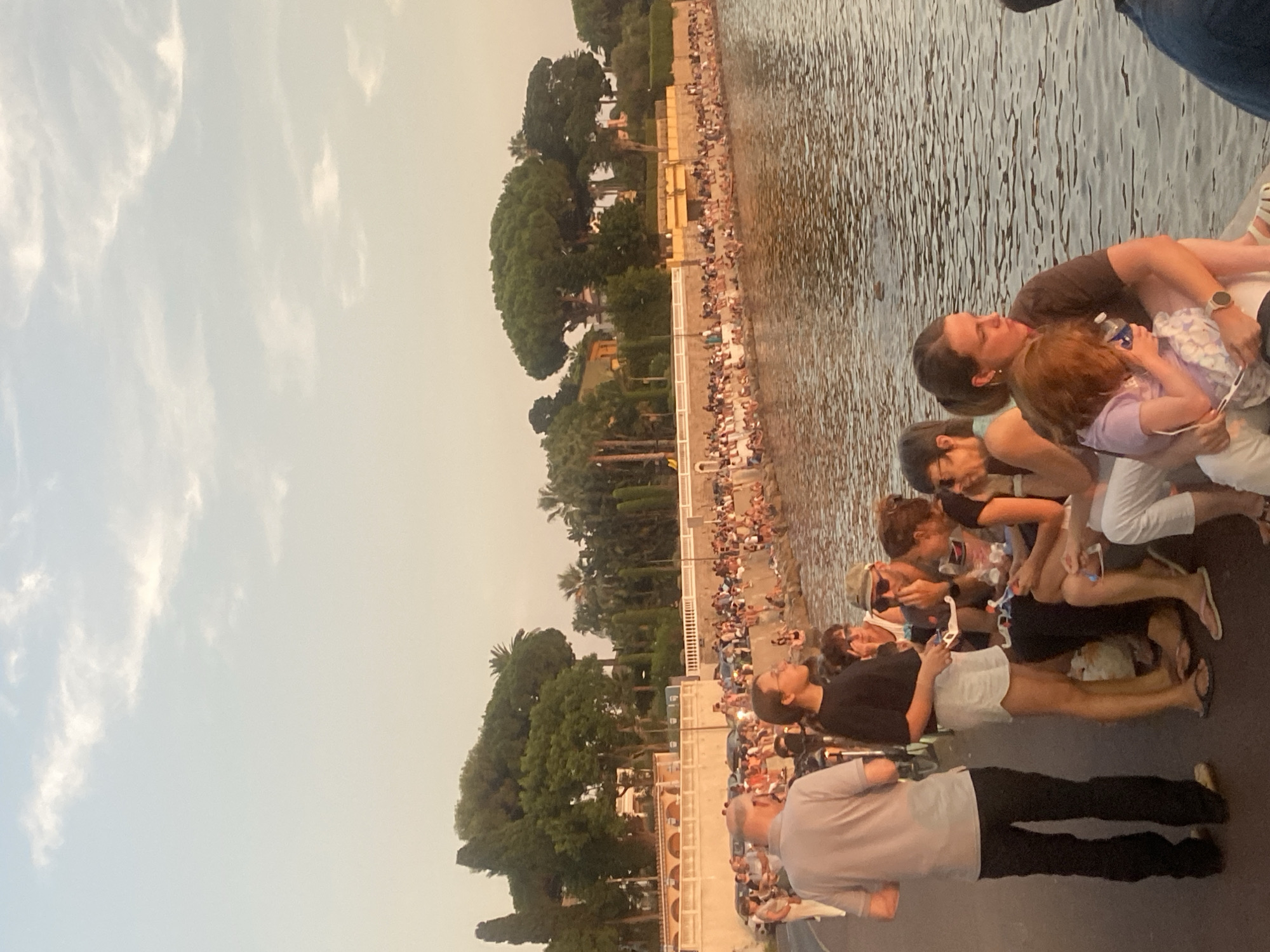 The image captures a scene along the Promenade du Port in Antibes, France, during what appears to be late afternoon or early evening. A crowd of people are gathered along the waterfront, some sitting on a low wall while others stand. The water in the foreground is calm and reflects the soft, warm light of the setting sun, creating a gentle orange hue. Behind the people, a street lined with parked cars is visible, suggesting the location is a popular public area. Further back, lush green trees and buildings with arched structures can be seen, typical of the Mediterranean architecture. The sky is a pale blue with scattered, wispy clouds. In the foreground, several individuals are in focus, including a man in a grey polo shirt and black pants, a woman in a black t-shirt and white shorts, and a woman holding a child. Some people are interacting with each other, while others are looking out at the water or the surrounding area. The atmosphere seems relaxed and communal.