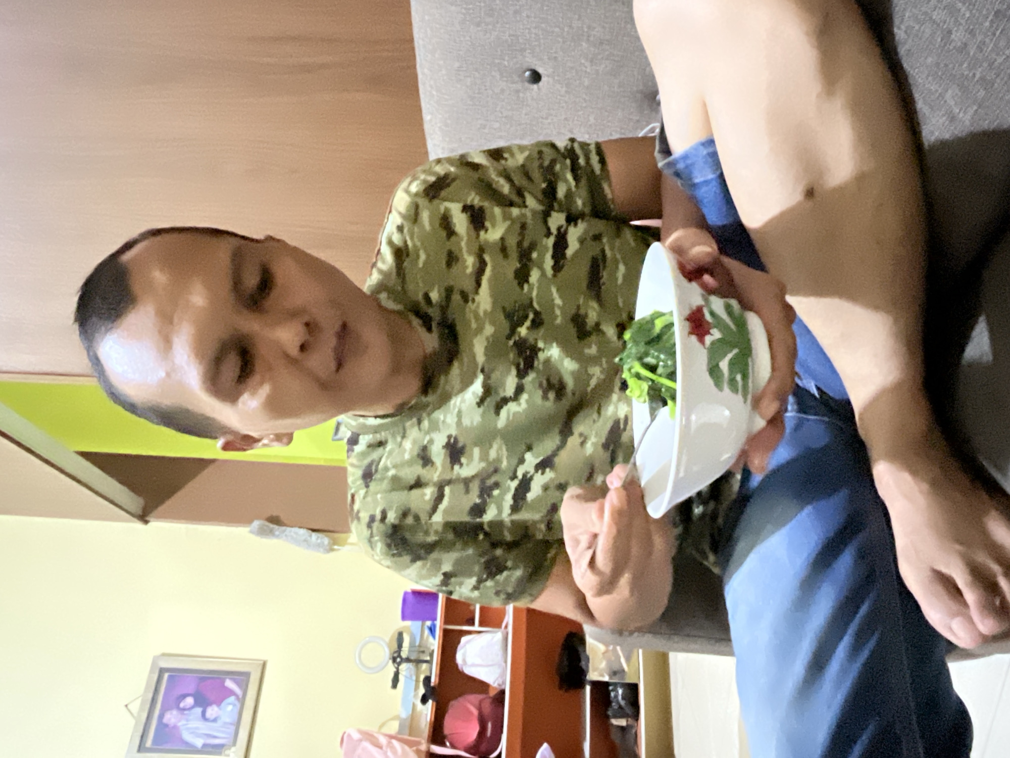 A person wearing a camouflage t-shirt and blue jeans is sitting on a gray couch, holding a white bowl with green leafy vegetables inside. The person's left leg is crossed over their right knee. The background includes a yellow wall with a framed picture, a brown shelf unit with various items, and a hint of a green ceiling. The location is likely a residence, but there is no information to determine if the location is in Lhokseumawe, Indonesia.