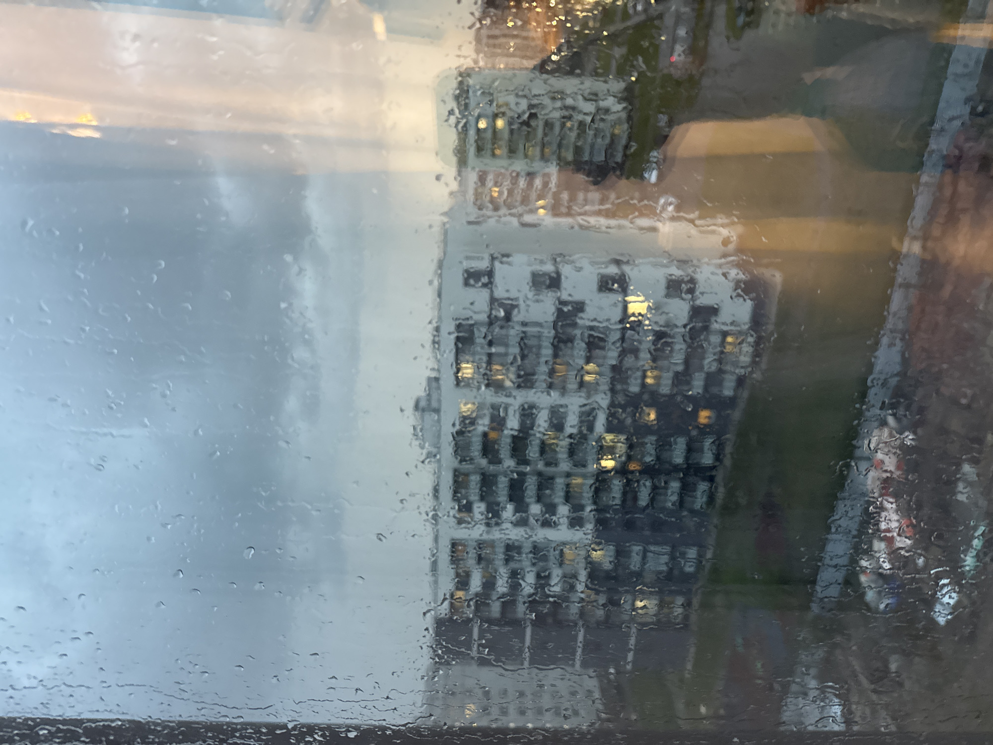 The image shows a rain-streaked window with a cityscape visible through it. The sky is overcast, and a multistory building with illuminated windows is partially visible. The scene is blurry and indistinct due to the raindrops on the glass. The location appears to be urban, but the specific city and exact details are unclear.