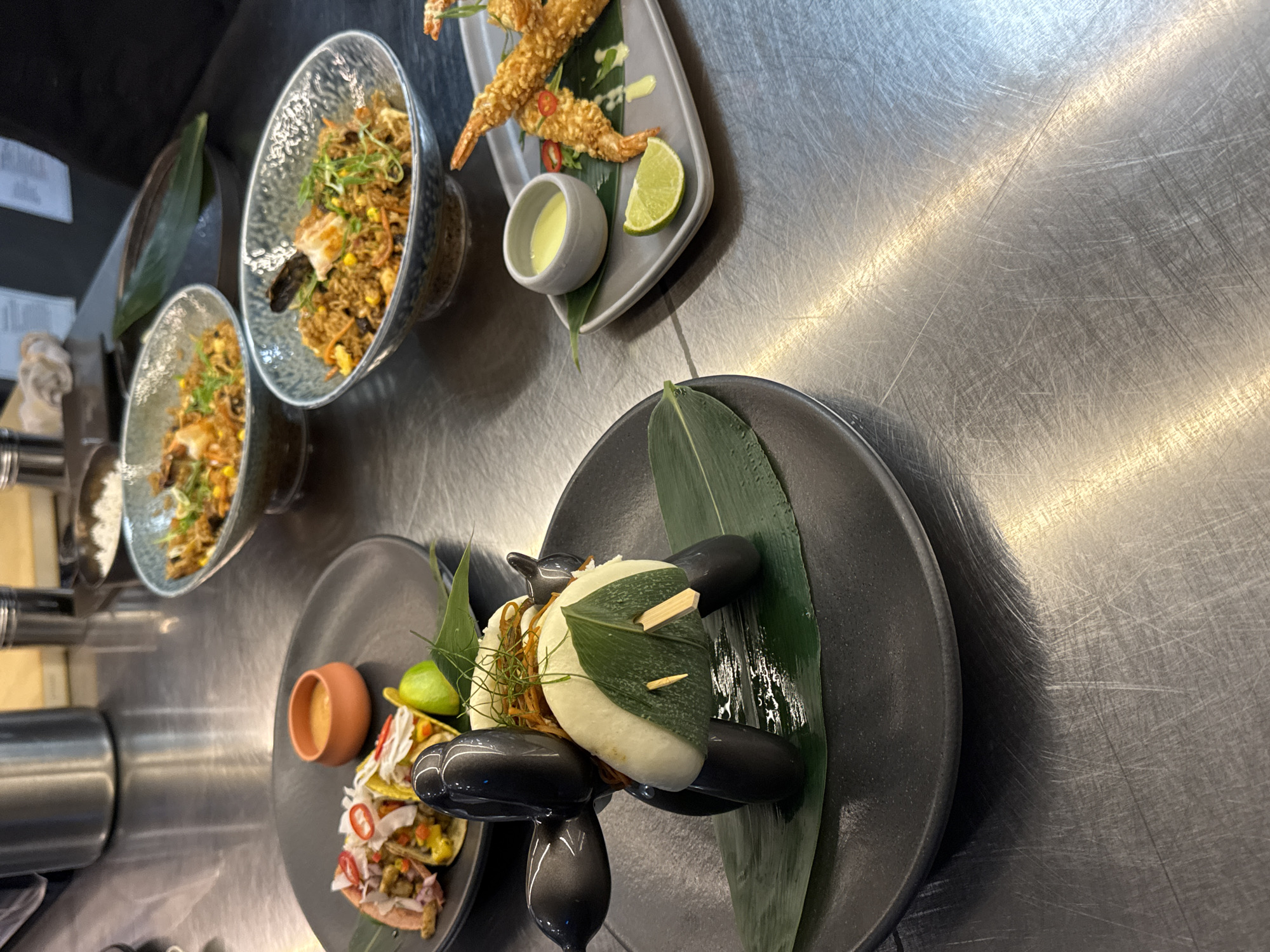 A still life food photograph displays an assortment of dishes artfully arranged on a stainless steel countertop, suggestive of a restaurant kitchen environment. The scene is lit from above, casting reflections and shadows on the metallic surface. In the foreground, a dark gray plate holds a stylized, dark gray balloon animal sculpture resembling a dog, with a steamed bun topping its back, garnished with shredded vegetables, a green leaf, and a wooden skewer. To the left, another dark plate features three mini tacos, a small terracotta dish filled with orange sauce, a lime wedge, and garnished with chili peppers and shredded vegetables. In the mid-ground, two bowls of what appears to be fried rice or a similar grain-based dish are visible. One bowl is closer to the viewer and contains a mix of rice, vegetables, and what looks like a piece of seafood, topped with green onions. Further back, a similar bowl holds the same type of dish. To the right, a rectangular gray plate holds three pieces of golden-fried shrimp, a lime wedge, and a small dish of pale yellow sauce, with a slice of red chili pepper visible. A single dark green leaf is placed beneath the shrimp. The background is slightly blurred, showing stainless steel canisters and what might be a kitchen appliance. The overall impression is one of culinary presentation and artistic plating. The image does not contain any information about the city of Sofia, Bulgaria.