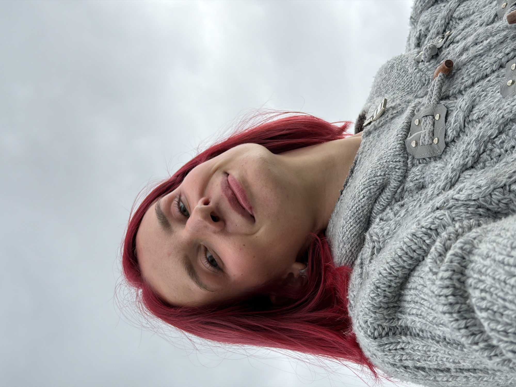 A female individual with vibrant red, shoulder-length hair is depicted in a low-angle shot, looking directly at the camera with a subtle smile. She wears a thick, light gray cable-knit sweater featuring distinctive gray leather-like toggle fastenings adorned with metal studs, and wooden toggles secured by rope loops. A metal zipper pull is visible near the collar. The background consists of a uniform, overcast light gray-blue sky. The scene is located in the city of Chaikovsky, Russia.