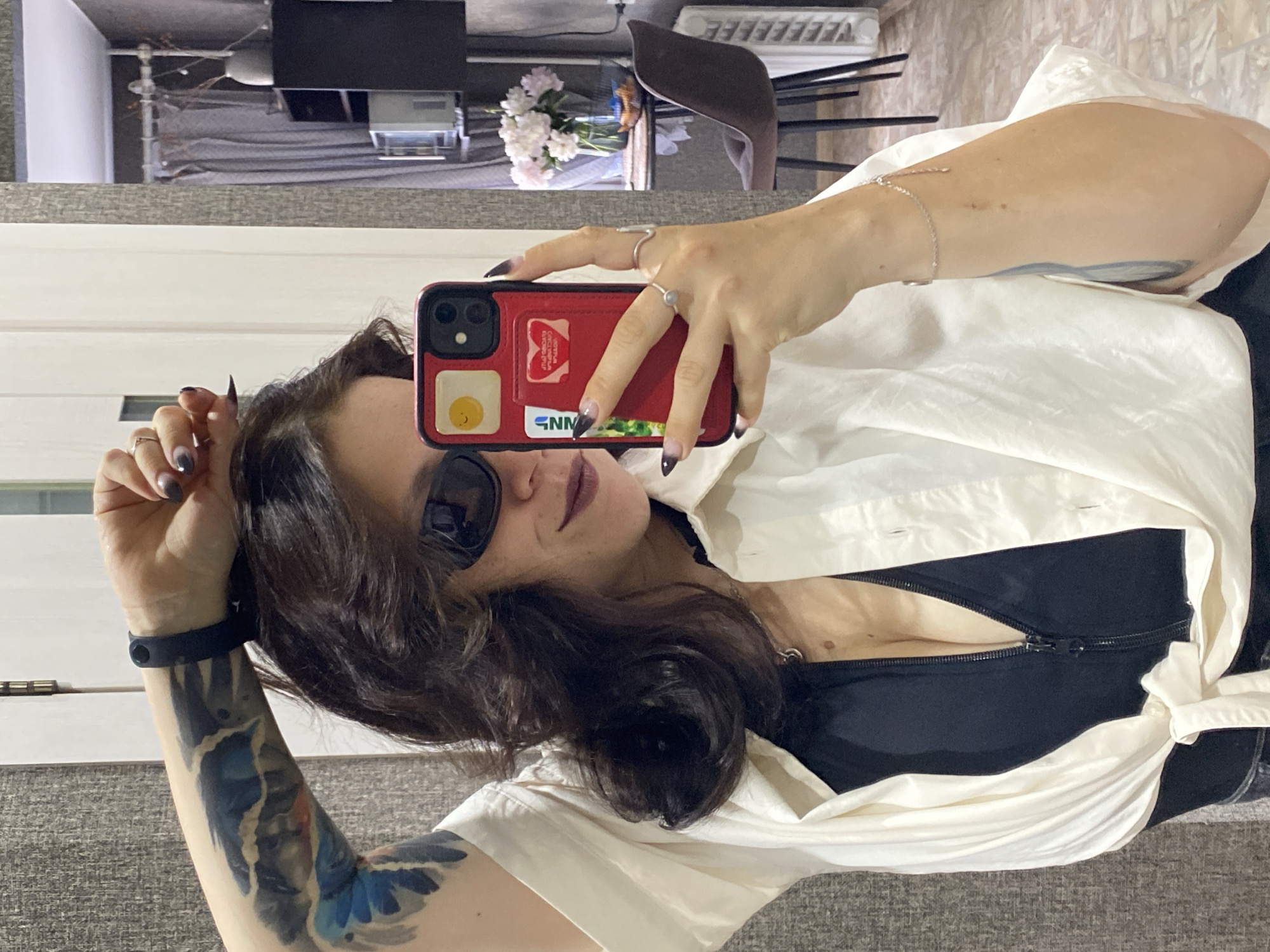 In an indoor setting, a woman is taking a mirror selfie. She is positioned on the left side of the frame, with her right arm raised and bent, her hand resting on her head. She is wearing a white unbuttoned shirt over a black top that is partially unzipped, revealing her cleavage. She has on dark sunglasses and dark lipstick. Her left arm is visible, adorned with a tattoo. On her right hand, she wears several rings, and a silver bracelet is on her right wrist. She holds a red phone with a yellow sticker of a smiley face and two cards in its case, in her right hand, at chest level, to capture the reflection. The background shows a wall with a textured pattern and a doorway to another room. In the reflection, a table with a vase of pink and white flowers, a chair, and a radiator are visible. The location is identified as Chaikovsky, Russia.