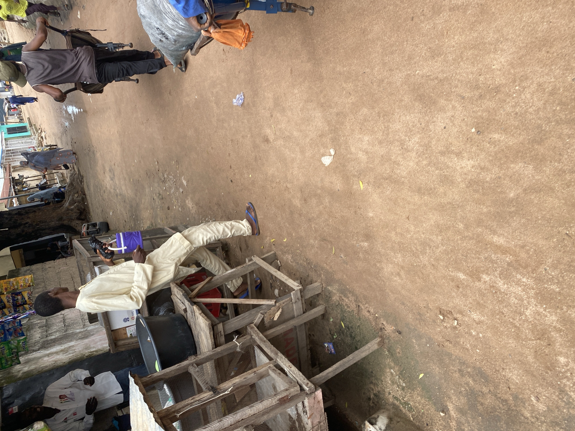 The image captures a scene in a market or street in Jalingo, Nigeria, characterized by its dusty, reddish-brown ground. In the foreground, a young man wearing a light-colored traditional outfit and blue flip-flops is leaning against a wooden stall. He is holding a small purple container in his right hand and a dark object, possibly a phone, in his left.
Behind him, a man in a white shirt with a portrait on it stands, appearing to interact with someone off-camera. To his left, a large black basin sits on the wooden stall.
Further in the background, several other individuals are visible. A man in a grey tank top and dark pants pushes a two-wheeled cart with a large sack on it. To his left, a woman in a patterned headscarf and grey dress walks away, pulling a cart. In the distance, more people and market stalls line the street, with buildings and vegetation visible. The overall impression is one of a bustling, open-air marketplace in an African city.