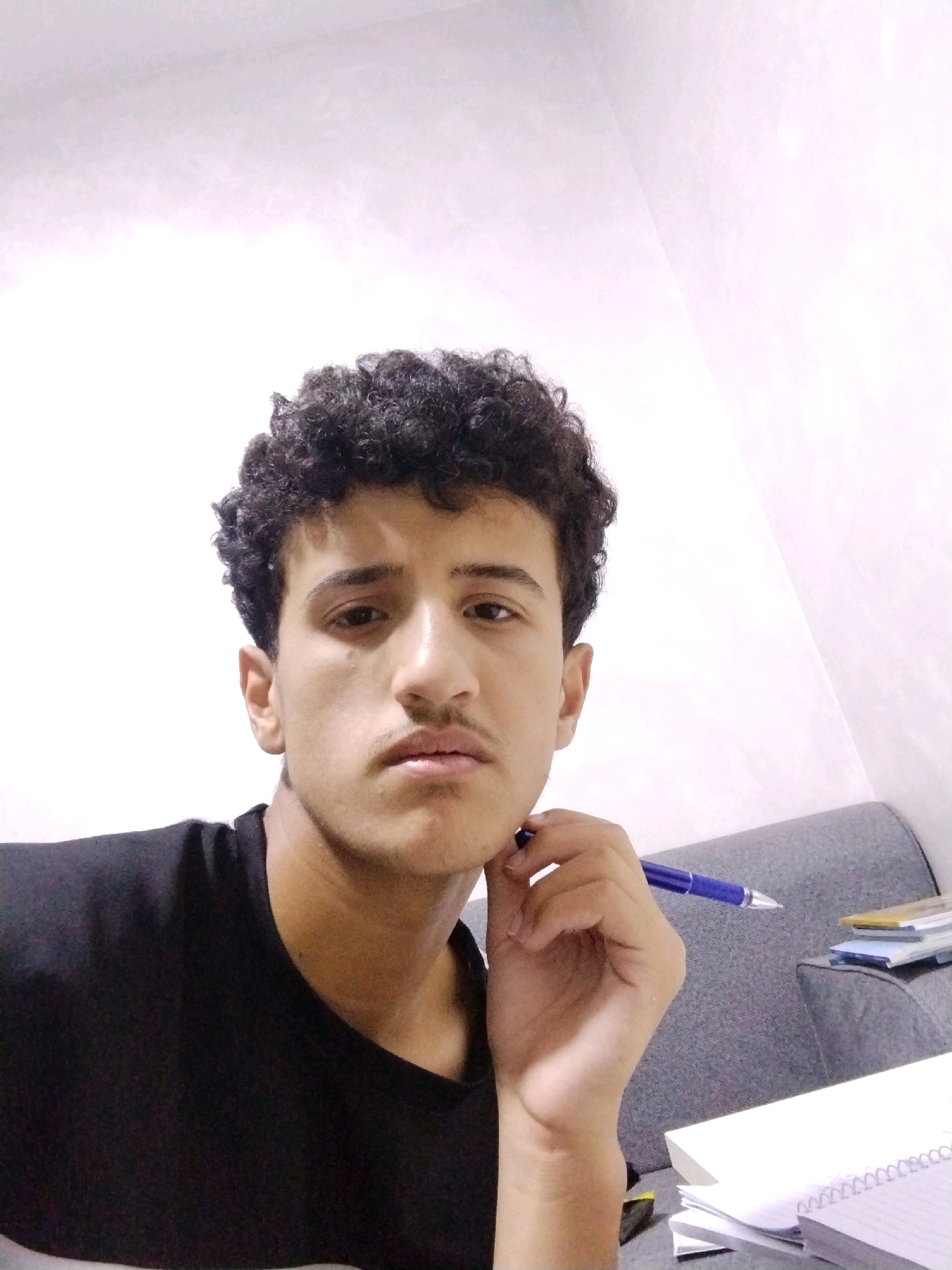 A young man with dark, curly hair and a light mustache is shown in a close-up selfie taken indoors. He is wearing a black t-shirt and resting his chin on his right hand, which is holding a blue pen. The background consists of a white wall with textured shadows and a gray upholstered surface, possibly a sofa, with a stack of books and notebooks on it. The setting appears to be a home or study area in Al-Hamra, Jordan.