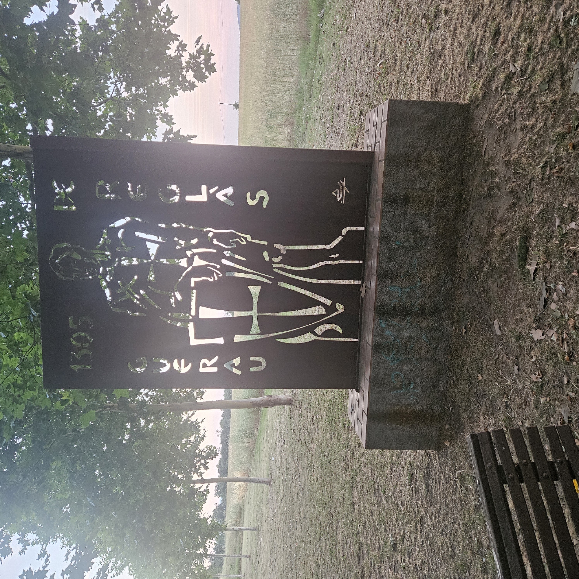 The image depicts an outdoor monument with cut-out lettering and a silhouette of a figure. The monument is dark-colored, likely metal, and stands on a brick and stone base. The cut-out text on the monument reads "1305 DE GUERAU DE ROCAFORT". The silhouette appears to be of a man holding a shield with a cross. To the left of the monument, a wooden park bench is partially visible. The environment surrounding the monument consists of dry grass, fallen leaves, and several trees with green foliage. In the background, a field of dry grass or crops extends to the horizon, with a pale sky above. The base of the monument has some graffiti. This monument is located in the municipality of Aiguaviva, in the province of Girona, Spain.