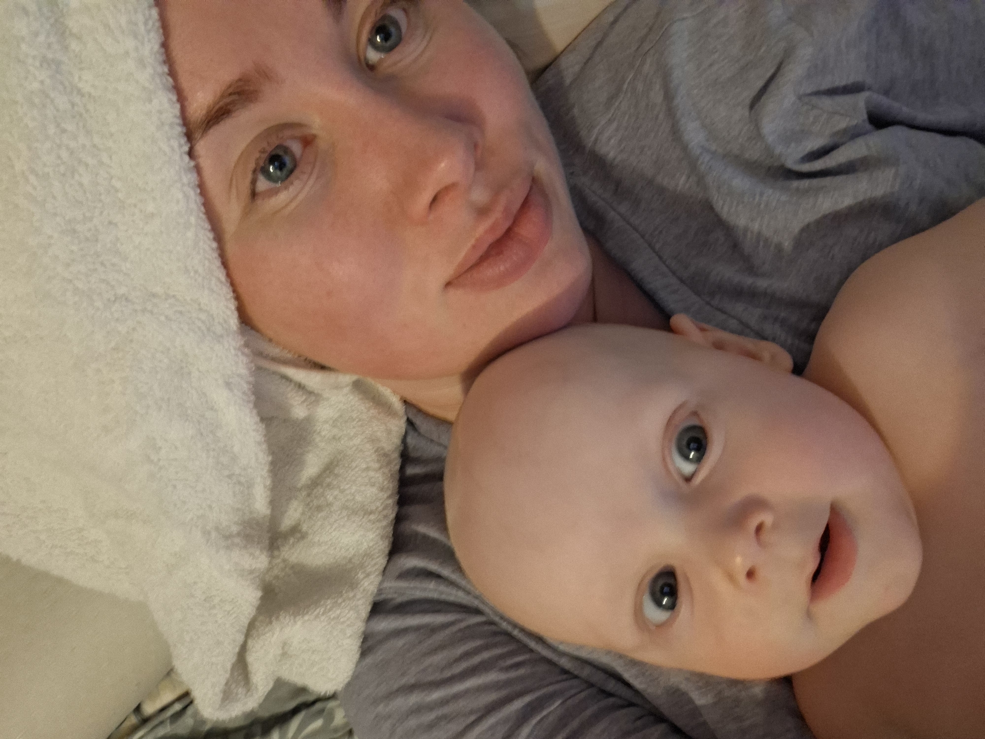 A close-up shot depicts a woman and a baby lying down together. The woman has fair skin, light eyes, and is wearing a gray shirt. A white towel is positioned behind her head. The baby, with a bald head and light eyes, lies close to the woman. The background appears to be a bed with a patterned sheet. The location is unknown.