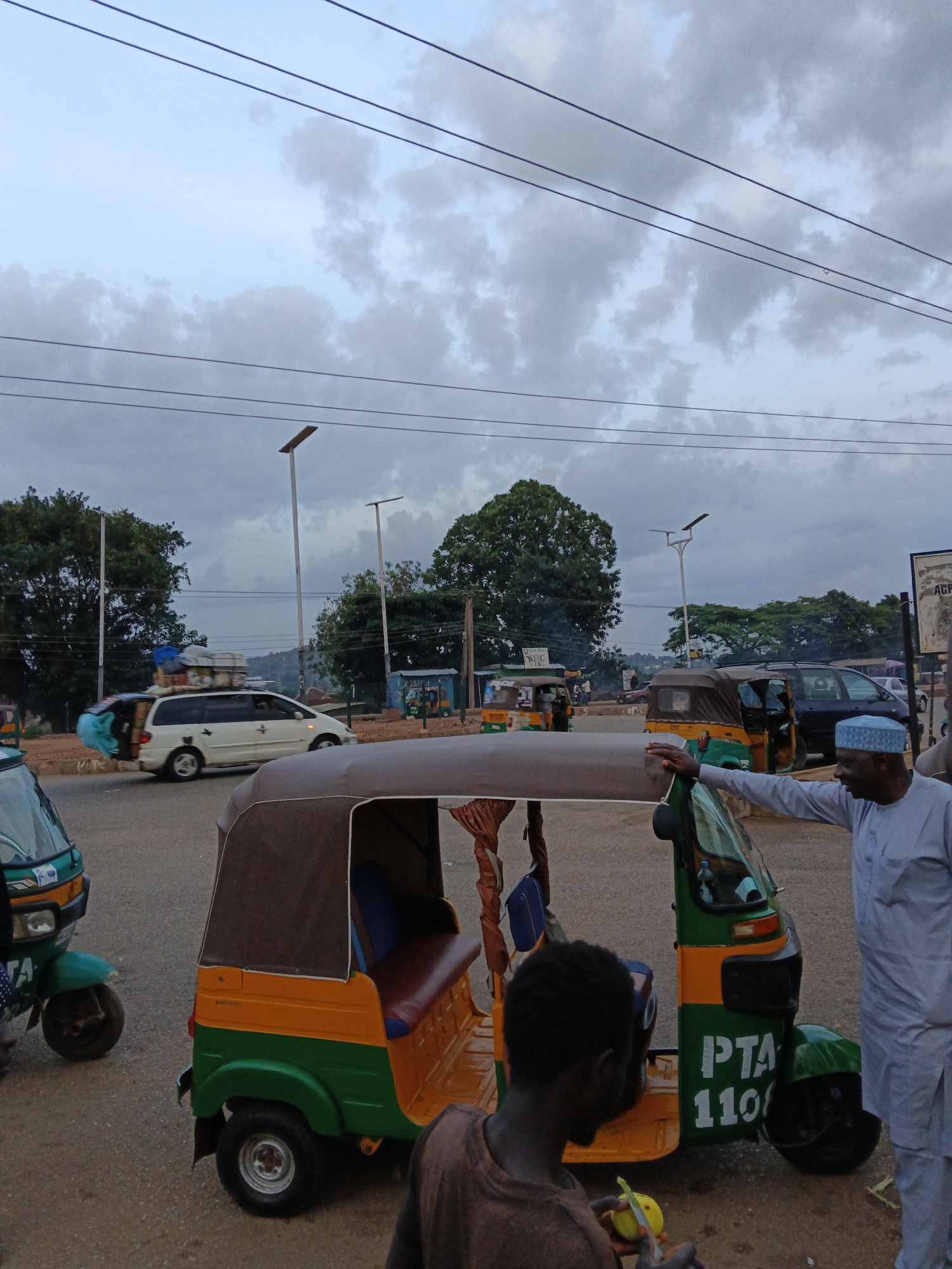In the city of Unknown, Unknown, under a cloudy sky, a street scene unfolds with multiple vehicles and people. In the foreground, a person is partially visible, facing away from the camera, holding a yellow object. To the right, a man in a light blue kaftan and matching cap stands near a green and yellow auto-rickshaw, with his right arm resting on its canopy. The auto-rickshaw has "PTA 110" written on its side. Behind this, another auto-rickshaw is visible. Further in the background, a white minivan overloaded with luggage is in motion, its roof and sides piled high with bags. Several trees dot the landscape, and two streetlights are present. The ground is paved with dirt and asphalt.