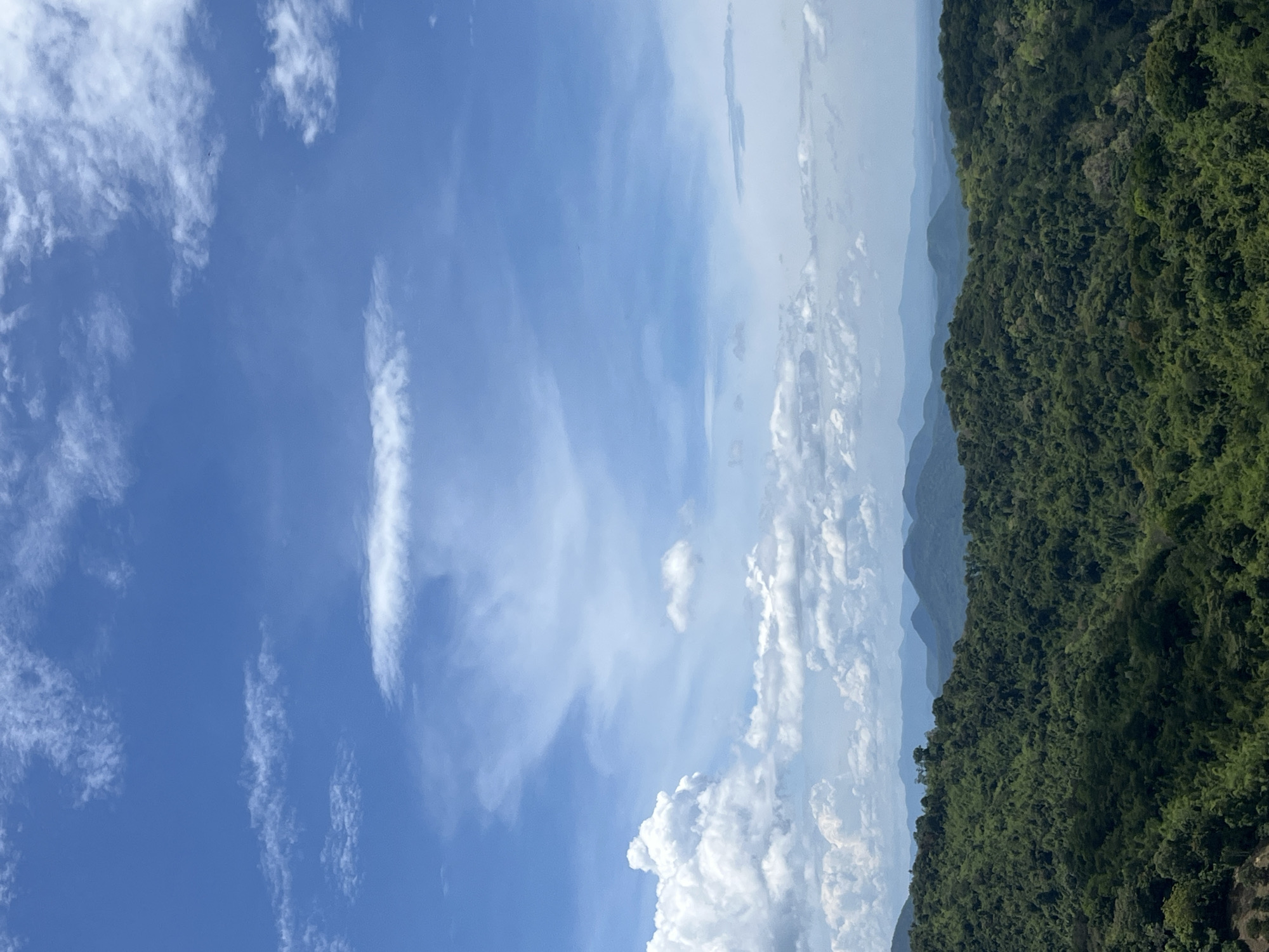 The image depicts a panoramic view of lush green mountains under a bright blue sky with scattered white clouds. The foreground is dominated by a dense forest covering rolling hills. In the background, several layers of mountains recede into the distance, becoming progressively bluer and hazier, suggesting a vast landscape. The sky is a vibrant blue with wispy white clouds, and larger, puffier clouds are visible on the left side, indicating fair weather. The scene appears to be a natural landscape, possibly a viewpoint overlooking a mountainous region. This specific landscape could be found in the rural or mountainous outskirts of Son Dien, Vietnam, known for its scenic natural beauty and hilly terrain.
