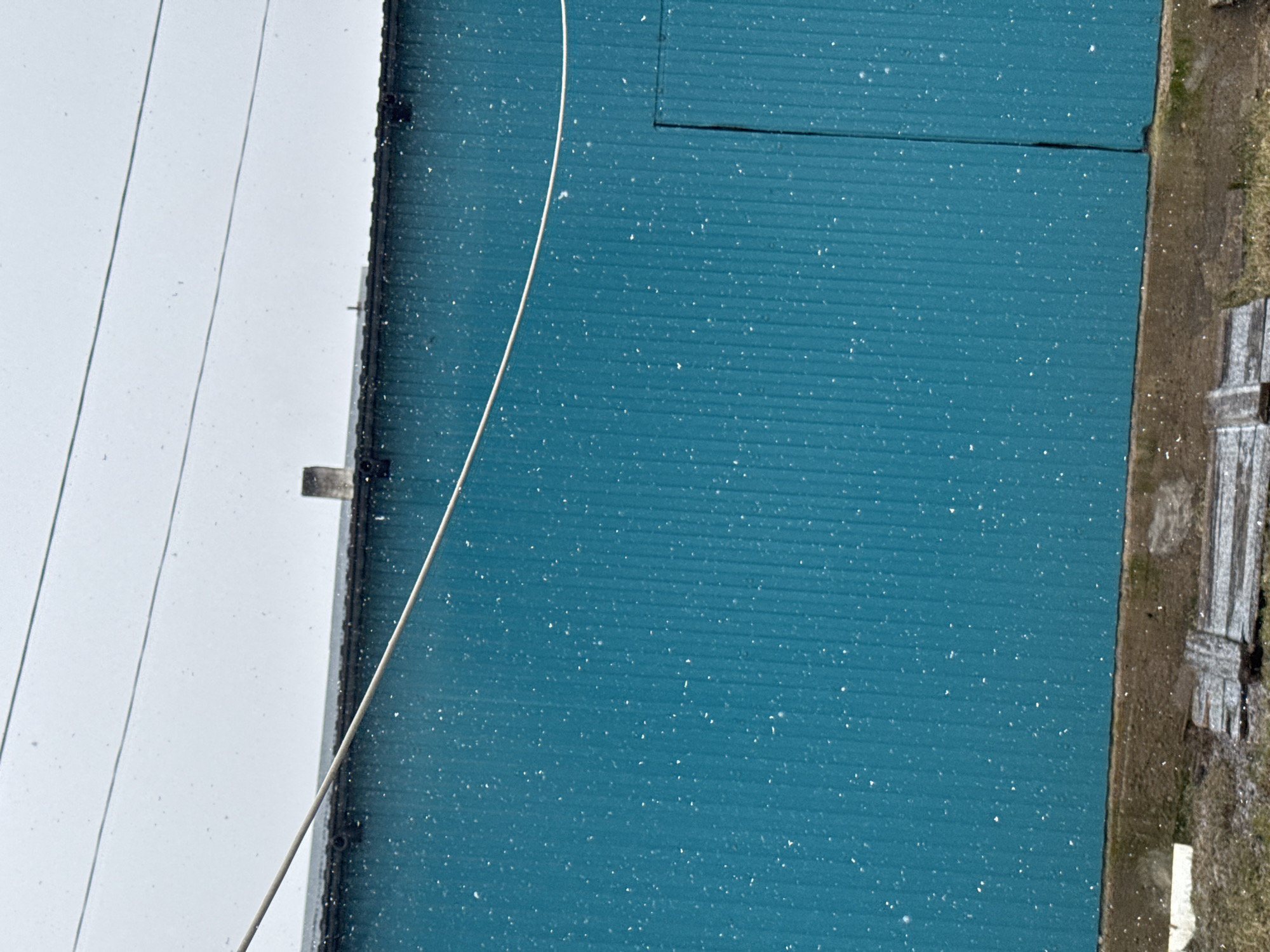 An exterior shot depicts a blue corrugated building wall with a door, partially covered in what appears to be snow. Next to the building is a stretch of land with old planks on it. A white cable runs across the wall. Adjacent to the building is a white surface, likely another wall or the sky, with power lines visible. The photograph is taken from a slightly elevated angle. The location is Уральское, Russia.