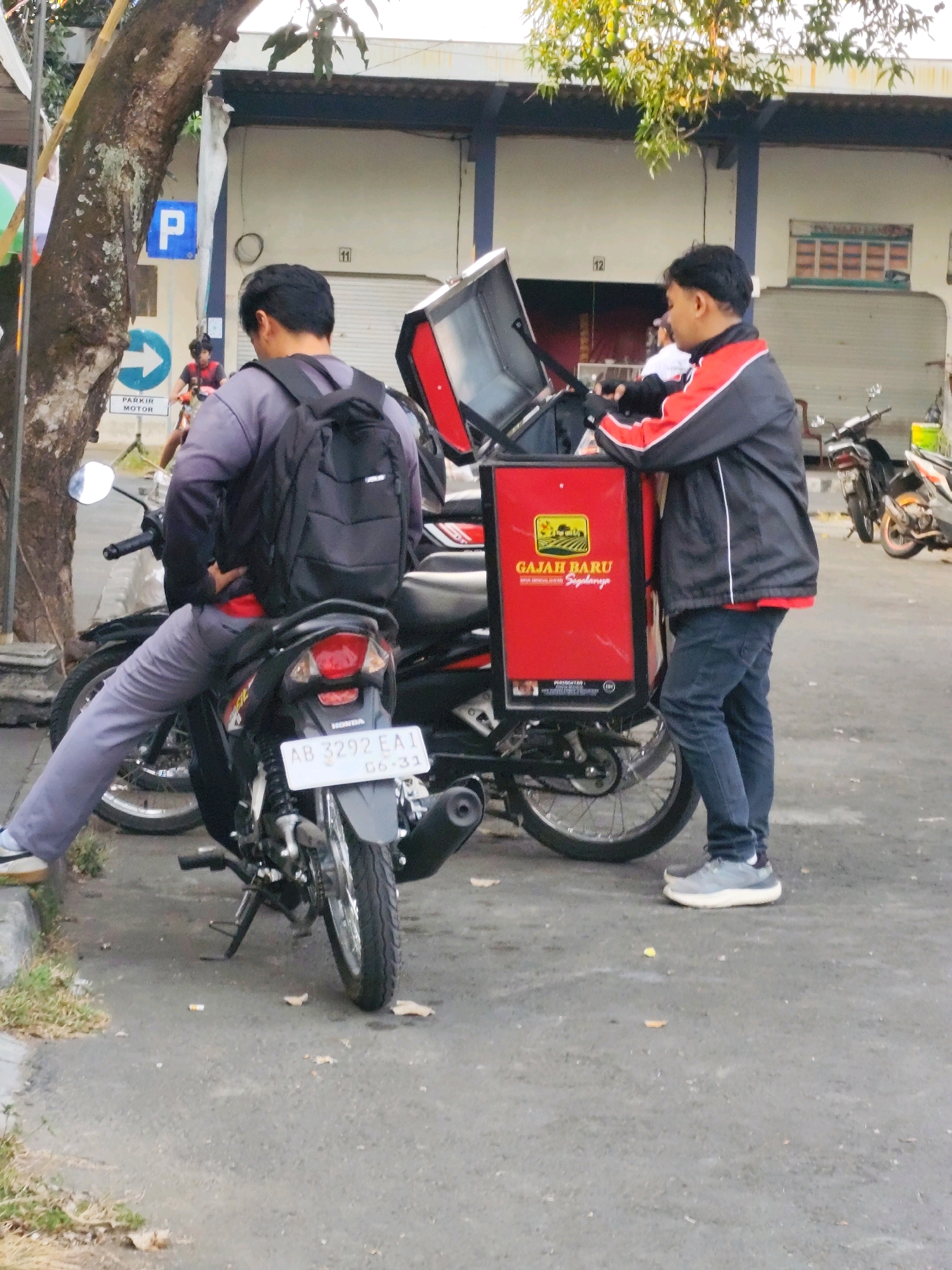 Two men stand next to motorcycles in what appears to be a parking area in Bantul, Indonesia. The man on the left, wearing a grey shirt and pants, is seated on a motorcycle and has a black backpack on. The man on the right, wearing a black jacket with red and white stripes over a red shirt and dark pants, is standing and appears to be interacting with a red delivery box attached to the back of a motorcycle. The delivery box has text and a logo on it. In the background, there is a building with a large roll-up door, and street signs. The scene is outdoors, with a tree on the left and pavement on the ground.