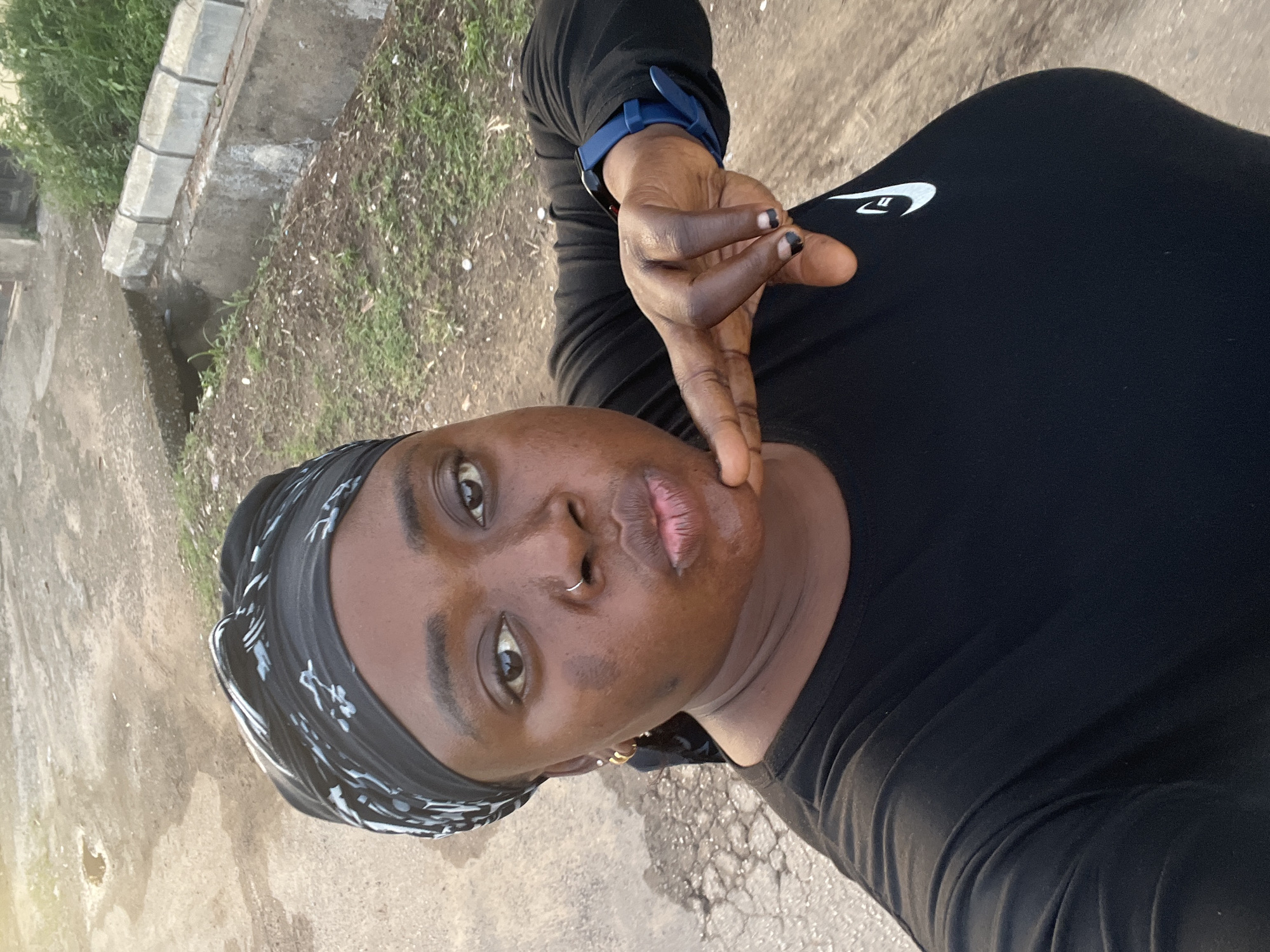 A person is taking a selfie outdoors. The person is wearing a black long-sleeved shirt with a white logo. They are also wearing a blue smartwatch on their left wrist and a black and white patterned headscarf. The person has a nose ring. The background consists of a sidewalk, curb, grass, and what appears to be a drainage ditch. Location cannot be determined.
