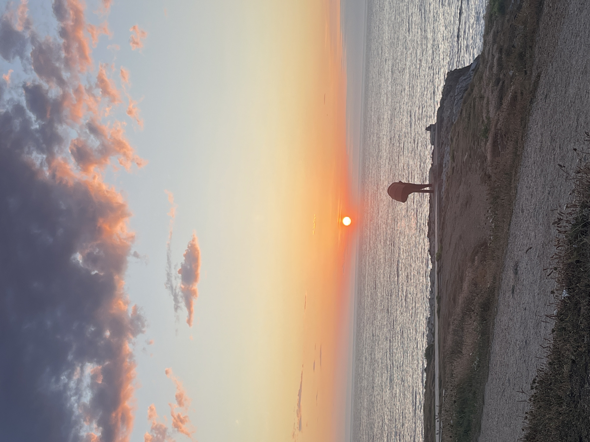 A silhouette of a person with their back to the camera stands on a cliff overlooking the ocean at sunset. The person is wearing a dark top and pants and appears to be bending over, perhaps looking at something on the ground. The sun is low on the horizon, casting a warm orange glow across the sky and water. The sky is filled with soft, wispy clouds tinged with pink and orange. The cliff is covered with dry grass and dirt, with a path winding down towards the viewer. The ocean is calm, with gentle waves lapping at the shore. The image appears to be taken in A Coruña, Spain, a city known for its beautiful coastline and sunsets.