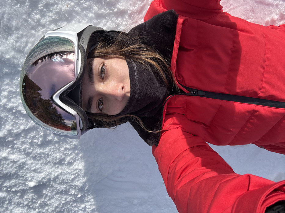A person lies on snow wearing a red ski jacket and a black neck gaiter. Ski goggles with a reflective lens rest on their head, reflecting trees and sky. The person looks at the camera with brown eyes. The snow is white with slight shadows. The jacket is zipped up and has a black zipper and black fur lining. The person's brown hair is visible.