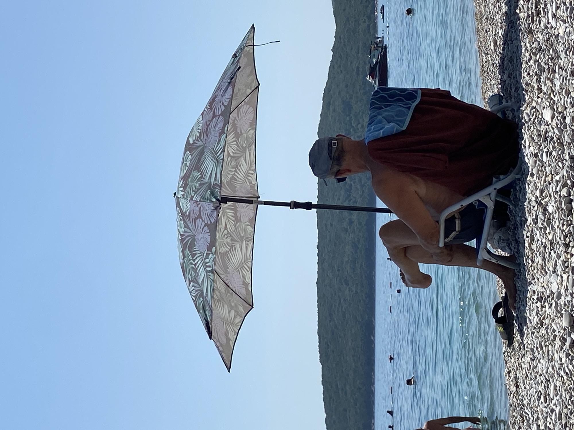A man is sitting in a beach chair under a patterned umbrella on a pebbled beach. The man is wearing a cap and sunglasses. A towel is draped over the back of his chair. The sea is calm with several people swimming and a jet ski in the distance. A forested hill forms the backdrop to the scene.
