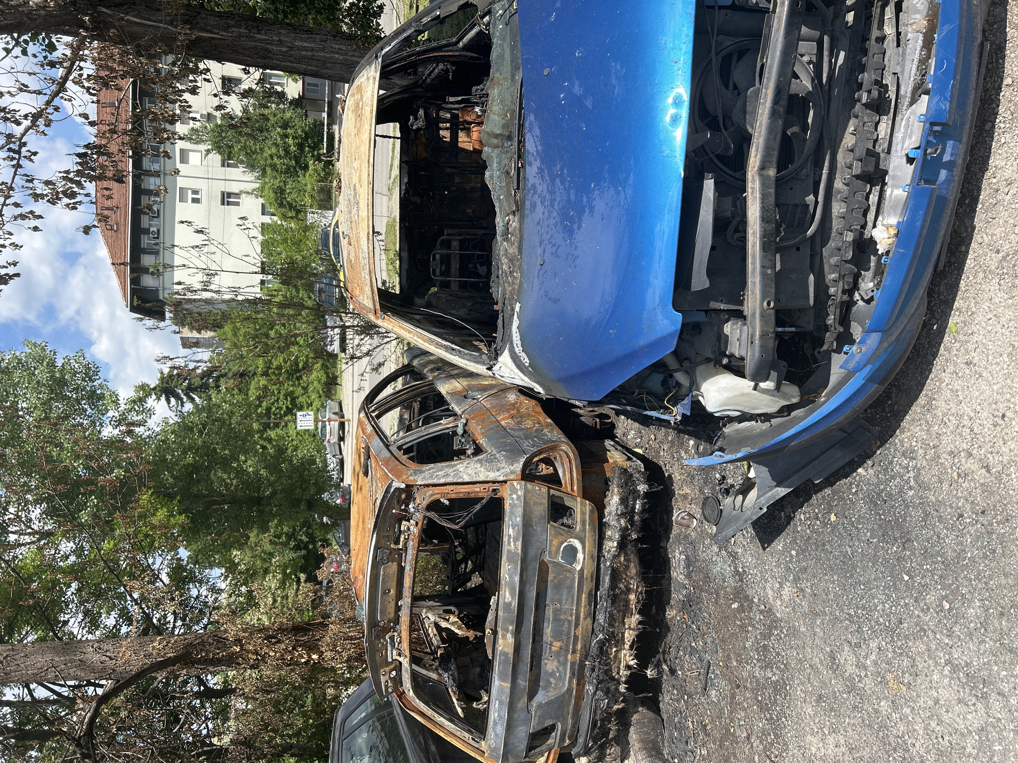 The image shows a severely burnt-out blue car on a street in Sofia, Bulgaria. The front end of the car, including the hood and engine compartment, is relatively intact and blue, while the rest of the vehicle is charred and gutted by fire. The vehicle’s interior is completely destroyed, with only remnants of the metal frame remaining. The car is parked on an asphalt road next to a tree. In the background, a light-colored building with a red-tiled roof is visible, suggesting an urban or suburban setting. Black soot and debris surround the car, indicating the intensity of the fire.