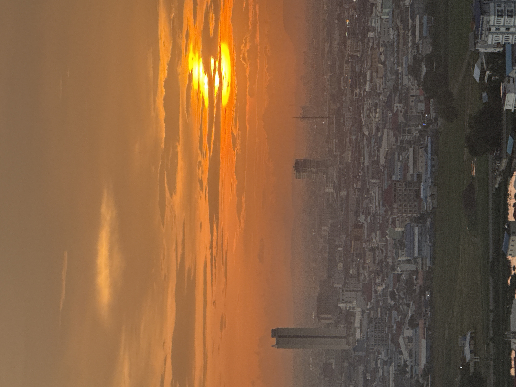The image depicts a hazy sunset over the city of Khan Mean Chey, Cambodia. The sky is dominated by vibrant orange and yellow hues, with the sun partially obscured by clouds. Below the sky, a dense urban landscape stretches out, characterized by a multitude of buildings of varying heights and architectural styles. A prominent, tall, rectangular building stands out on the left side of the frame, likely a skyscraper or office tower. In the mid-ground, other smaller buildings and structures are visible, interspersed with patches of greenery, possibly parks or undeveloped land. The overall impression is one of an expansive cityscape bathed in the warm, diffused light of the setting sun. No people are discernible in the image, and the focus is on the urban environment and the atmospheric conditions of the sunset.