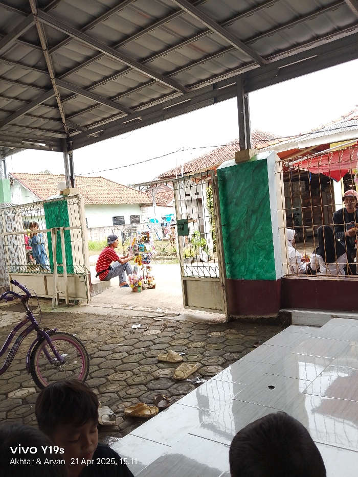 The scene takes place in the Kecamatan Anjatan area of Indonesia. The scene is a partially covered walkway with a metal roof and metal gates at the front. There is a bicycle parked on the left, and the viewer is looking towards the back of the walkway. There are several people in the scene, including a man sitting on the ground near a table with items for sale, a man sitting in the background through a gated area wearing a white shirt and pants, and a man through a gate with a red shirt and pants. There is also a man sitting in the background through the gate wearing black clothing and a white cap. There are a few other people obscured by the gate. There are several pairs of sandals lying on the ground of the walkway, possibly belonging to the people in the scene.