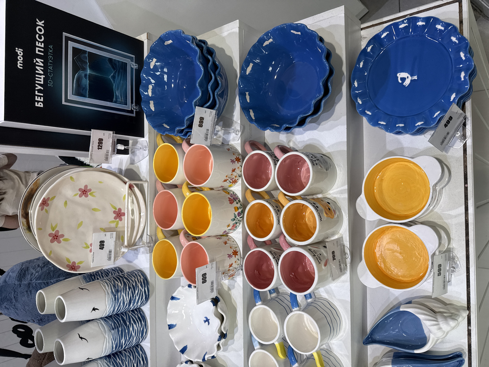 The image shows a display of ceramic tableware in a retail setting, likely a store located in Moscow, Russia. The items are arranged vertically on shelves, featuring a variety of colorful plates, bowls, and mugs. The objects are arranged in a grid-like pattern, with items stacked on top of one another. Each item is marked with a price tag. Specific items include blue plates with scalloped edges and animal motifs, mugs with orange and pink interiors, plates with floral designs, and vases decorated with a blue and white wave pattern and bird silhouettes. At the top left, a box labeled "БЕГУЩИЙ ПЕСOK" (Running Sand) with the brand name "modi" and the description "3D-СТАТУЭТКА" (3D-Statuette) is visible.