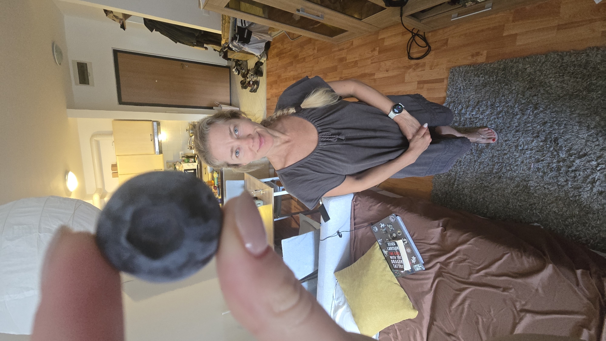 An indoor, portrait-style photograph, rotated 90 degrees clockwise, captures a woman holding a large blueberry in the foreground.
The main subject is a woman with blonde hair, styled in a braid, smiling faintly and looking directly at the camera. She has blue eyes and is wearing a dark grey, short-sleeved dress with a gathered neckline. A white smartwatch is visible on her left wrist, and her bare feet, with red nail polish on the toes, rest on a grey shaggy rug.
In the immediate foreground, a large, dark blueberry is held between fingers, one of which has light pink nail polish.
The background shows a home interior with light wooden flooring and a grey shaggy rug. To the woman's right, a brown couch or bed is visible, adorned with a yellow throw pillow. On the couch, a book is open, clearly displaying the title "STIEG LARSSON" and "THE GIRL WITH THE DRAGON TATTOO". Behind the woman, there is a light-colored chair and a table. Further back, a kitchen area with light wooden cabinets and a countertop is visible. Other elements include a wooden cabinet with glass doors, a white paper lantern, and a dark brown door with a black frame. Some clothes and shoes are seen on the floor near the wooden cabinet. The lighting indicates it is daytime, suggesting a casual, relaxed atmosphere in what appears to be a living room or studio apartment.