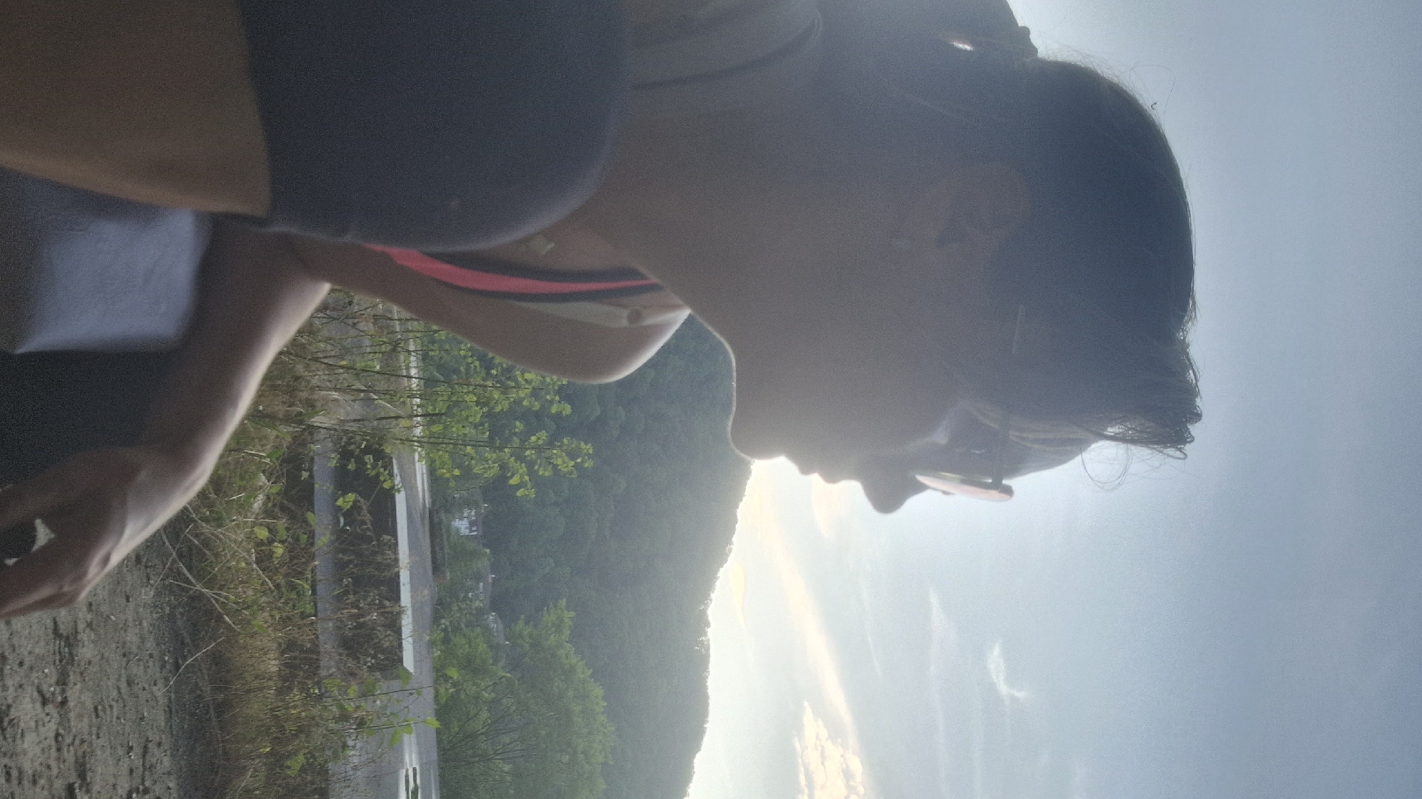 A person, seen in profile and from the shoulders up, is sitting outdoors against a backdrop of a hazy, sunlit sky and a distant, tree-covered mountain range. The person has dark hair pulled back and is wearing sunglasses. They are wearing a pink and black sports bra and dark shorts. The person is looking to the right, away from the camera. The background features a concrete structure, dry vegetation, and some greenery. The lighting suggests it is either early morning or late afternoon. No specific landmarks or context suggest the location is Sochi, Russia.
