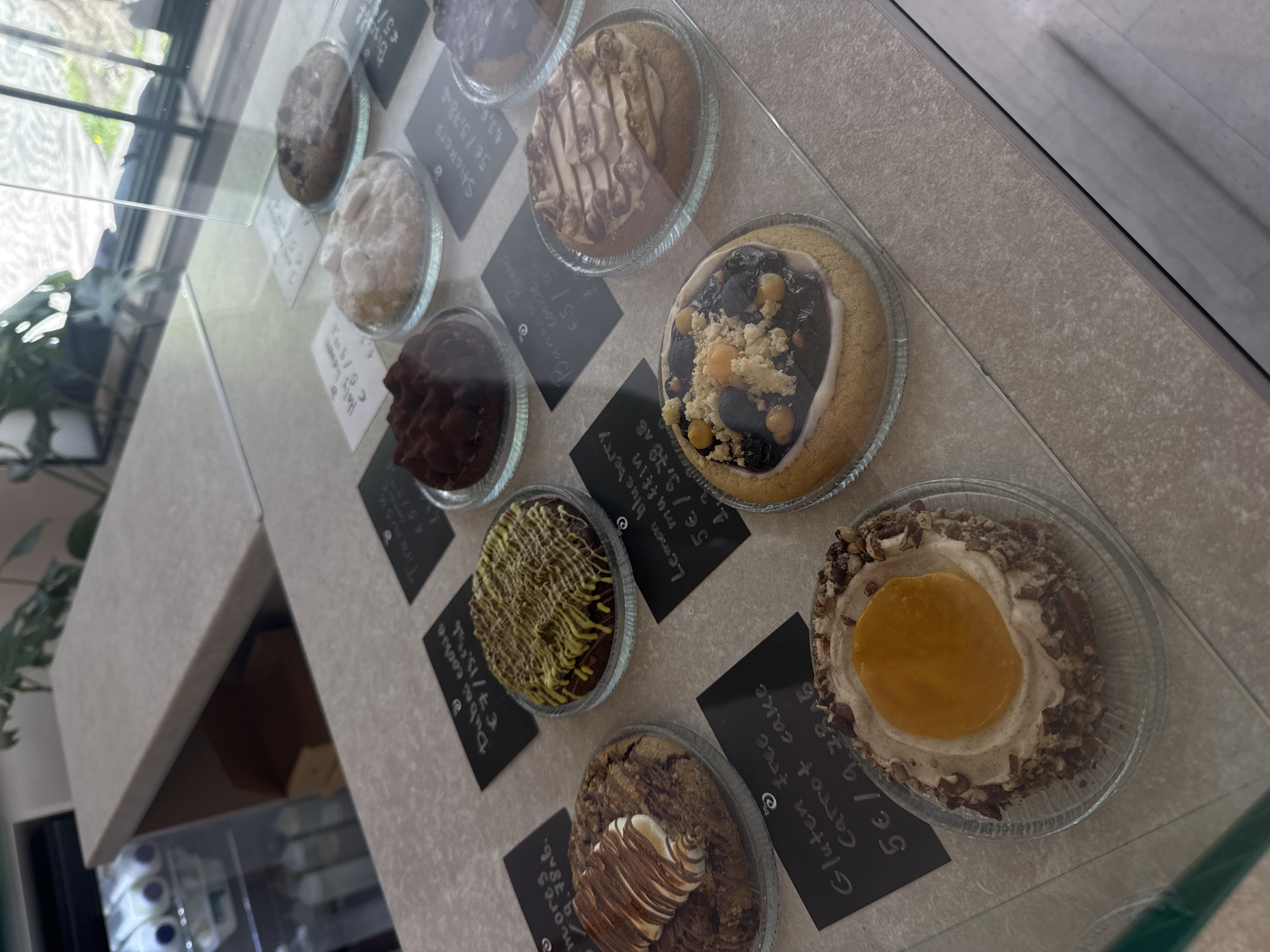 This image captures a close-up, slightly overhead view of a selection of baked goods displayed in a glass case, likely in a cafe or bakery in Sofia, Bulgaria. The main subjects are various desserts, each presented in individual clear glass or plastic dishes, arranged on a light-colored countertop.
The desserts showcase a variety of toppings and styles:
- One appears to be a "Lemon blueberry muffin" with a generous blueberry and crumble topping.
- Another is a "Gluten free Carrot cake" with a creamy, light-colored frosting, a circular orange/peach-colored center, and a perimeter of chopped nuts.
- A "Dubai cookie" features a dark base with intricate yellow icing drizzle.
- A "Tiramisu" is dark brown with a white creamy topping.
- A "Banana Bread cookie" is topped with white frosting and chocolate drizzle.
- Other items include "Stickers," "Holy Lewers," "Biscal," and "m-ores," though their specific appearance is less clear.
Each dessert has a small, black sign with white handwritten text indicating its name and price. Prices are generally in euros, such as "5€" or "7€", with some items listing two prices, e.g., "5€ / 9.78€". The glass display case reflects the indoor environment, including a window and some greenery outside, suggesting natural light. The setting is clean and well-lit, designed to present the desserts appealingly to customers.