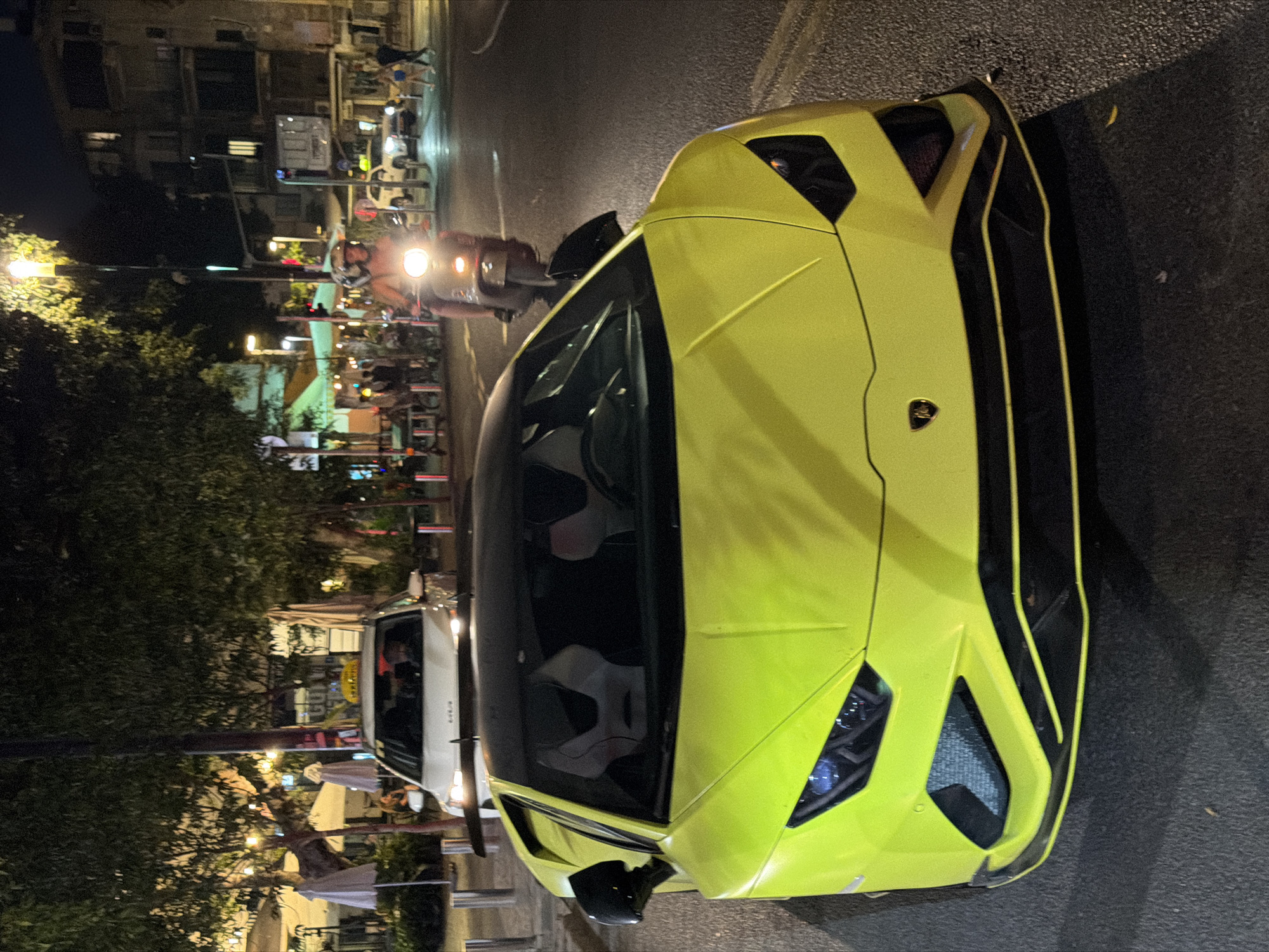 An overhead, low-angle shot captures a bright yellow Lamborghini supercar at night on a paved street in Tel-Aviv, Israel. The vehicle, a Lamborghini Huracán EVO, is positioned centrally, facing directly towards the viewer. It features a black roof, black side mirrors, and a distinct black front bumper with yellow accent lines that complement its neon yellow body. The interior reveals light-colored racing seats.
Behind the Lamborghini, a white Kia-brand taxi with its characteristic yellow roof sign is partially visible. Further in the background, a motor scooter with two helmeted riders approaches from the right side of the frame, its headlight brightly illuminated. To the far right, another white SUV is visible, along with several pedestrians.
The urban environment is dimly lit by streetlights, vehicle headlights, and the warm glow from multiple outdoor cafes and restaurants lining the street on both sides. Numerous trees with sparse foliage define the background, intermingled with building facades. White and beige umbrellas from outdoor seating areas are visible under the trees. Metal bollards delineate the roadside. The overall scene depicts a moderately active street at night in an urban setting.
