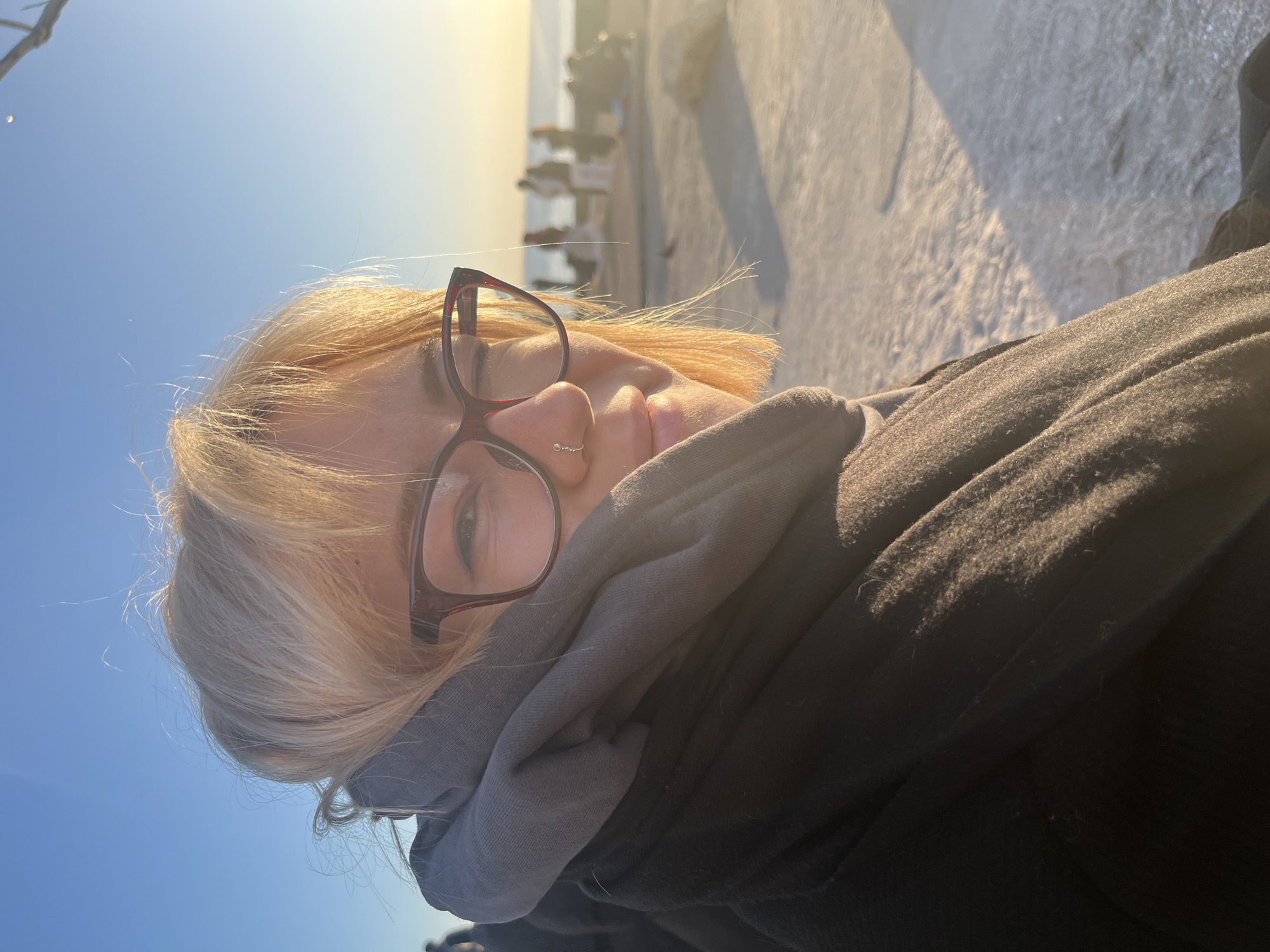 A blonde woman with glasses and a nose ring is bundled in a gray scarf and black jacket. She is outdoors on a concrete promenade with other people visible in the background. The location is likely Odesa, Ukraine, based on its coastal setting. The sun is shining brightly, creating harsh shadows. The sky is clear blue.