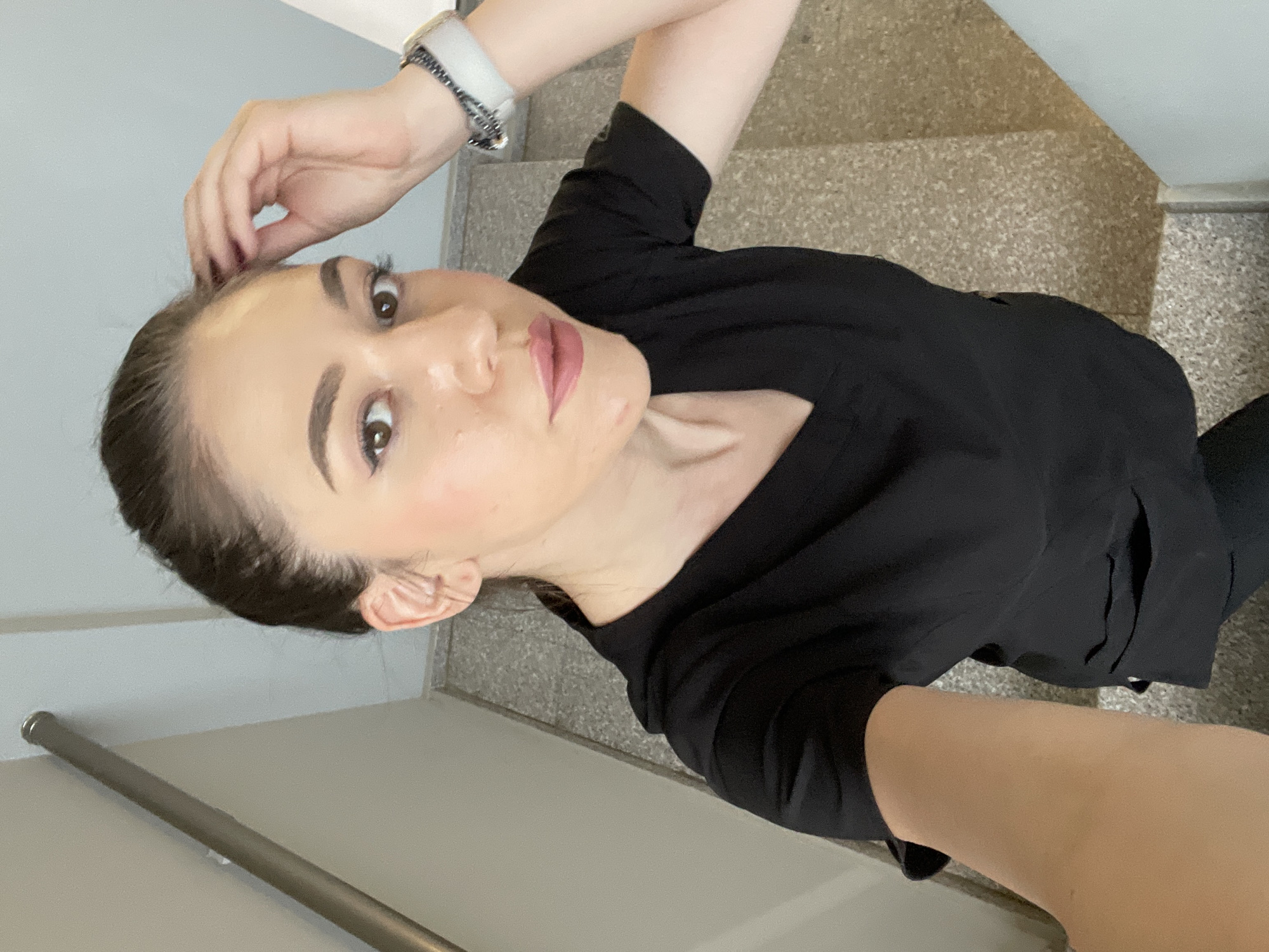 A woman is standing on a staircase, presumably in Sofia, Bulgaria, and is taking a selfie. She is wearing a black shirt and a watch on her left wrist. Her hair is pulled back, and she is looking directly at the camera. The staircase has speckled stone treads. There is a metal handrail running along the wall.