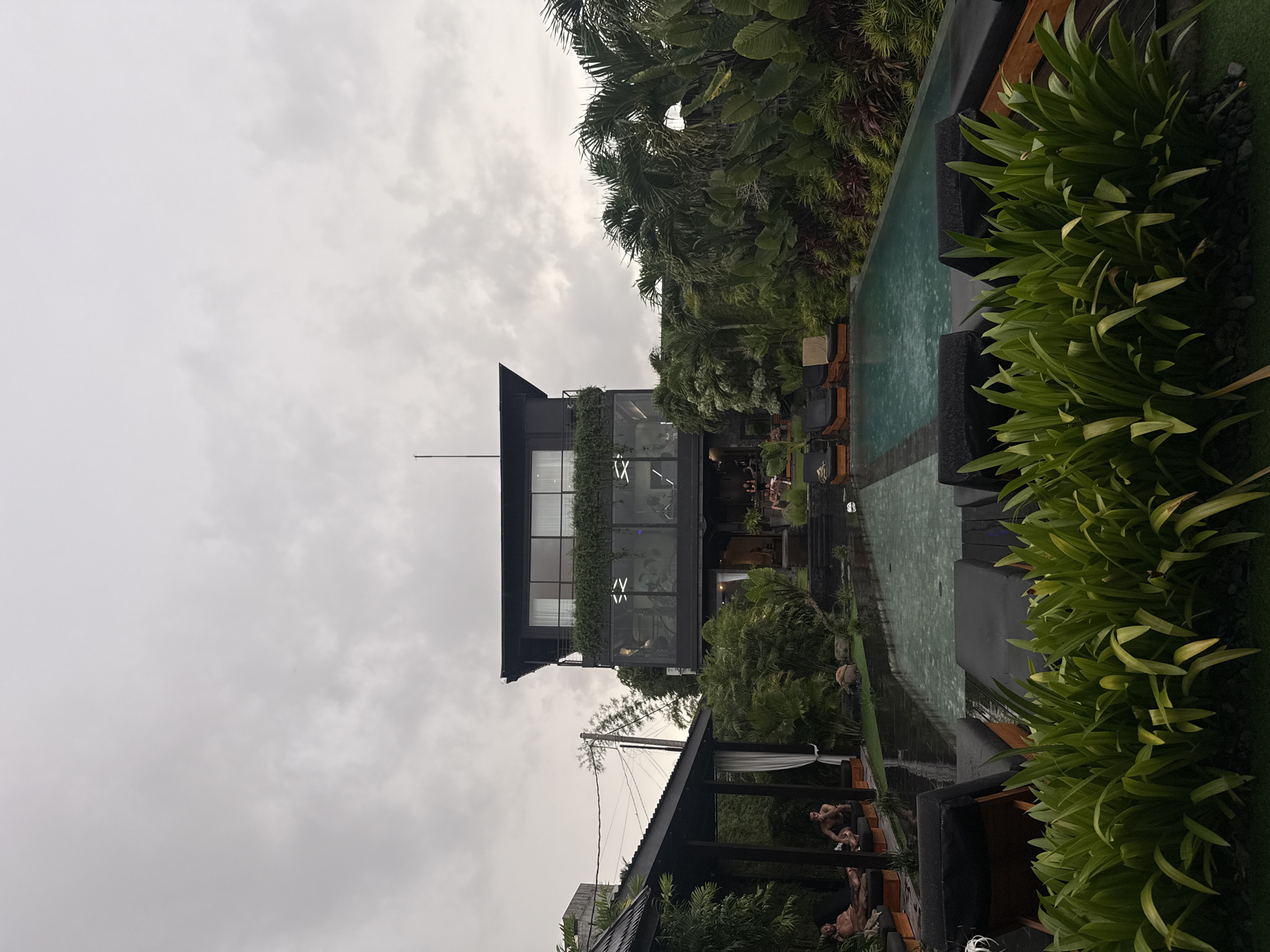 This media file captures an outdoor leisure or wellness area under an overcast sky.
**Subjects:** The scene features a modern, multi-story building with a dark exterior and large glass windows, positioned on the left side of the frame. The upper floor appears to be a fitness center or gym, with exercise equipment and figures visible through the glass. A planter box with lush green foliage runs along the base of the upper-level windows. To the right of the main building, a large swimming pool with greenish water stretches towards the background. The pool is bordered by dark-colored lounge chairs with cushions. Abundant tropical greenery, including palm trees and various plants, frames the pool area on the right and extends into the background. In the lower-left foreground, a smaller structure with a dark roof and white curtains is visible, beneath which several people are lounging. A utility pole with power lines is seen between the foreground structure and the main building.
**Activity or event taking place:** Several people are present, either relaxing on lounge chairs by the pool, sitting or lying down under the foreground structure, or exercising inside the glass-fronted building. The atmosphere suggests a leisure or wellness activity.
**Notable details:** The time of day appears to be during daylight hours, but the sky is completely overcast with thick grey clouds, resulting in dim, diffused lighting. The overall aesthetic is modern and lush, with dark architectural elements contrasting with vibrant green foliage. The water in the pool shows minor ripples, possibly from light rain or wind.
**Visible text:** Inside the upper floor of the main building, through the large windows, there are some illuminated white shapes that resemble stylized letters or symbols, possibly "X" or "IV". A small, unreadable logo or emblem might be present on one of the white sun loungers by the pool.